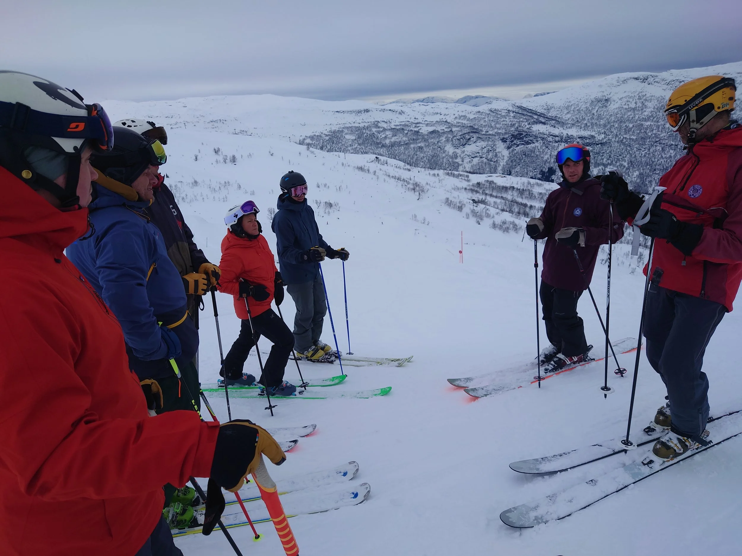 Gruppeskiløpere som står på snø med snødekte fjell i bakgrunnen.