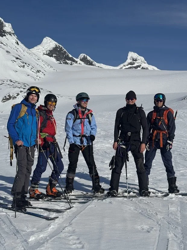 Fem personer i skibekledning står på snø med fjell i bakgrunnen i et snødekt terreng under klar blå himmel.