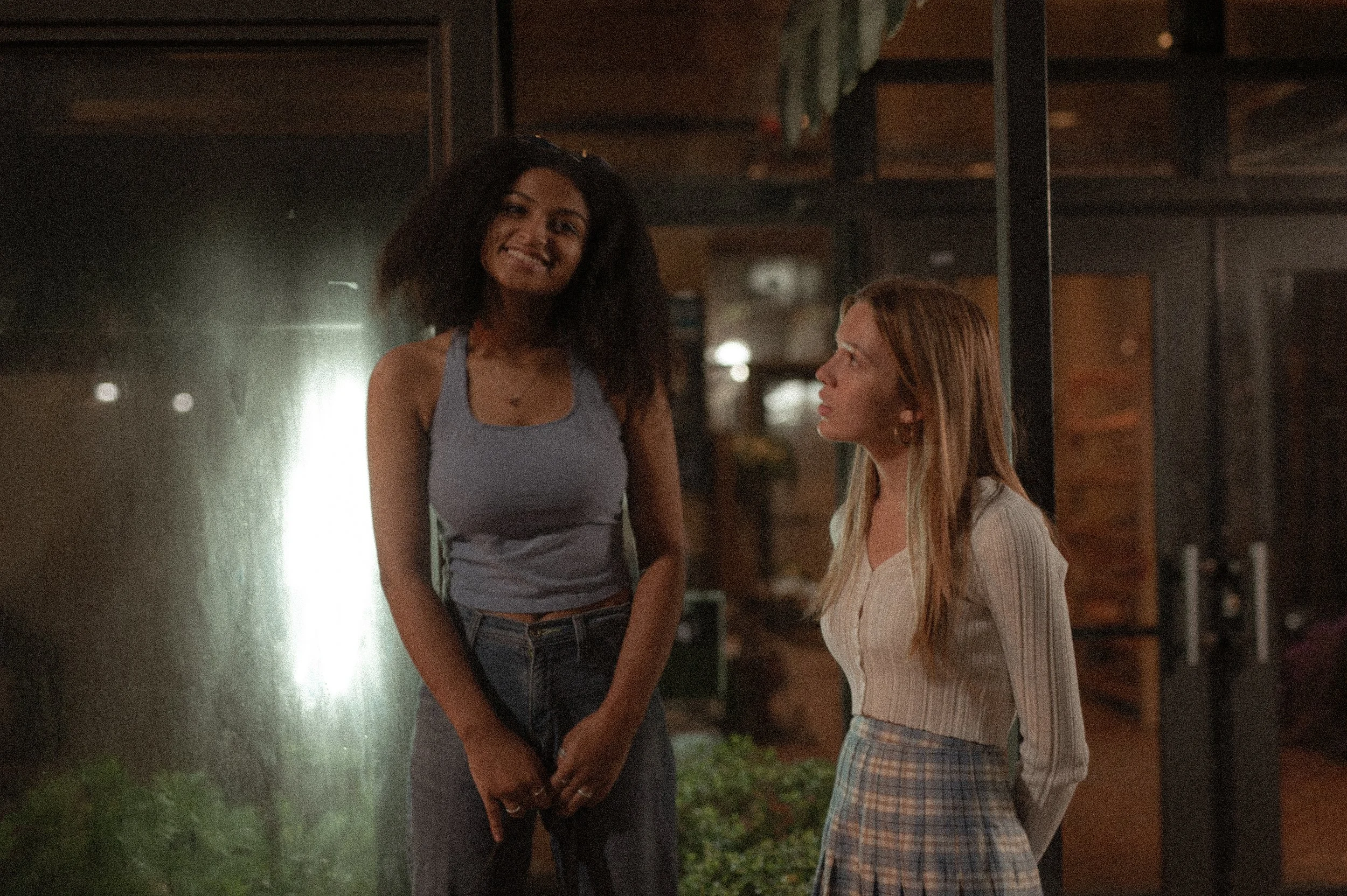 Two women standing outside at night, one with curly dark hair wearing a gray tank top and high-waisted dark jeans, smiling and showing teeth, the other with straight blonde hair wearing a long-sleeve beige top and plaid skirt, looking at the first woman