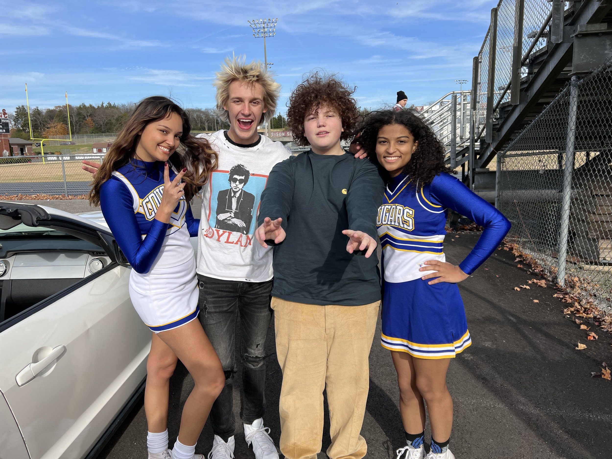 Four teenagers, two cheerleaders and two friends, posing together on a track field with a sports stadium and a car in the background, under a partly cloudy sky.