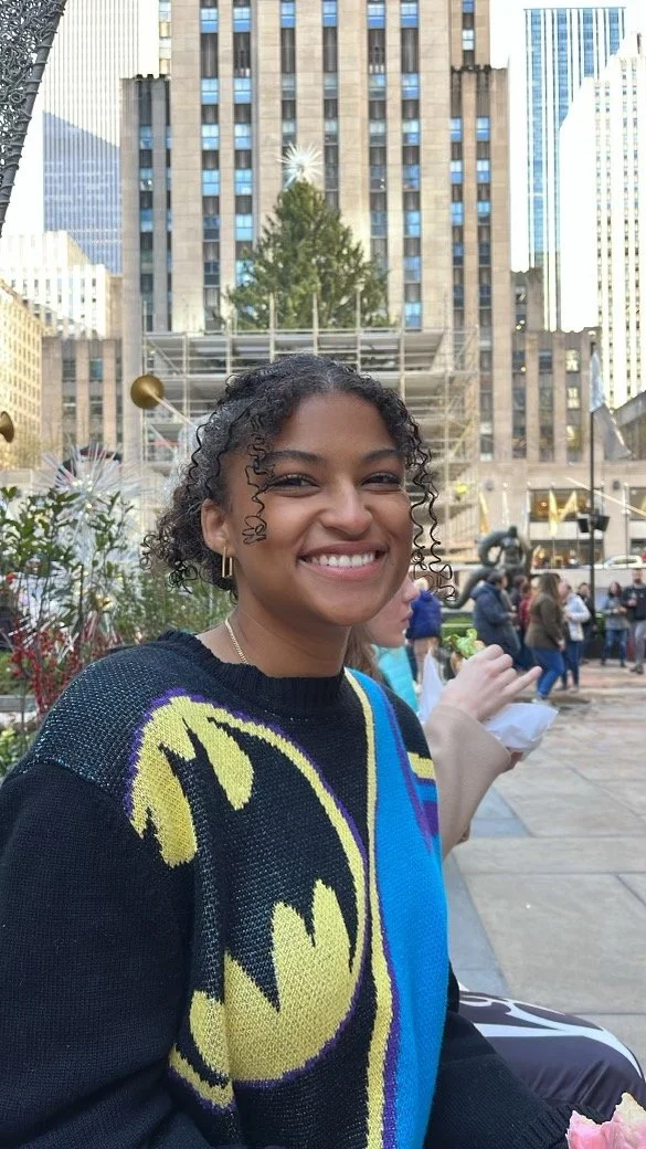 A young woman with curly hair smiling outdoors in a city plaza with tall buildings in the background, wearing a Batman sweater.