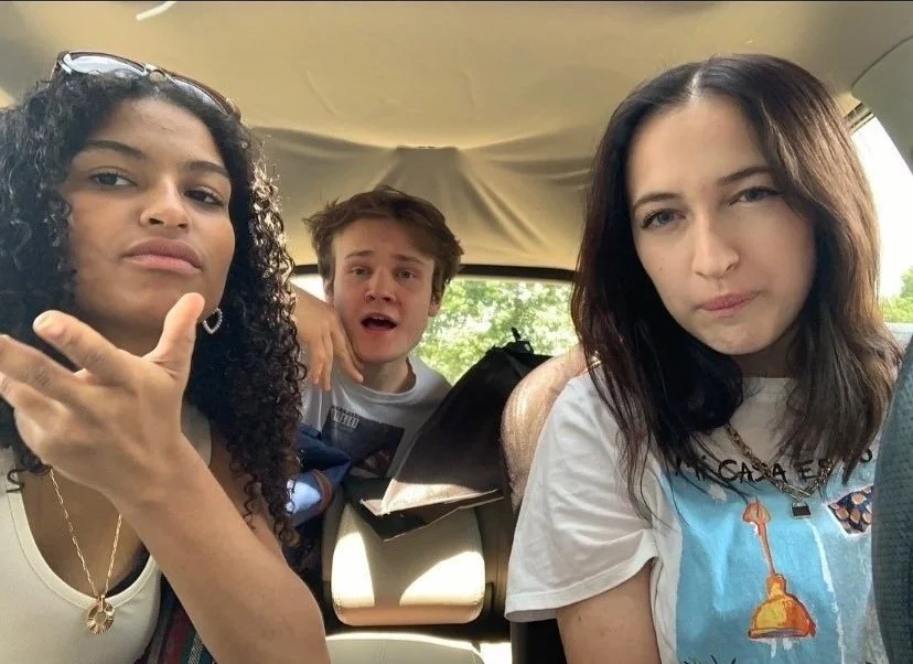 Three young people sitting inside a vehicle, making playful expressions, with the background showing trees and sky through windows.