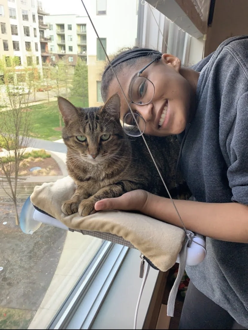 Person smiling and lying next to a tabby cat on a window perch, looking out a large window at an apartment complex and green outdoor area.