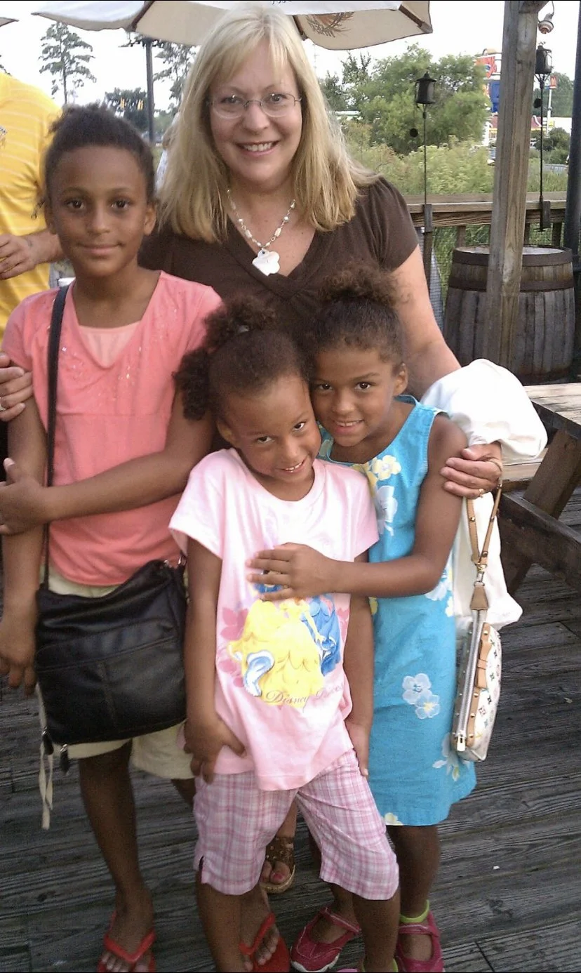 A woman with blonde hair, glasses, and a brown shirt is smiling and posing with four young girls on a wooden outdoor deck. The girls are wearing colorful dresses and are hugging each other and the woman.