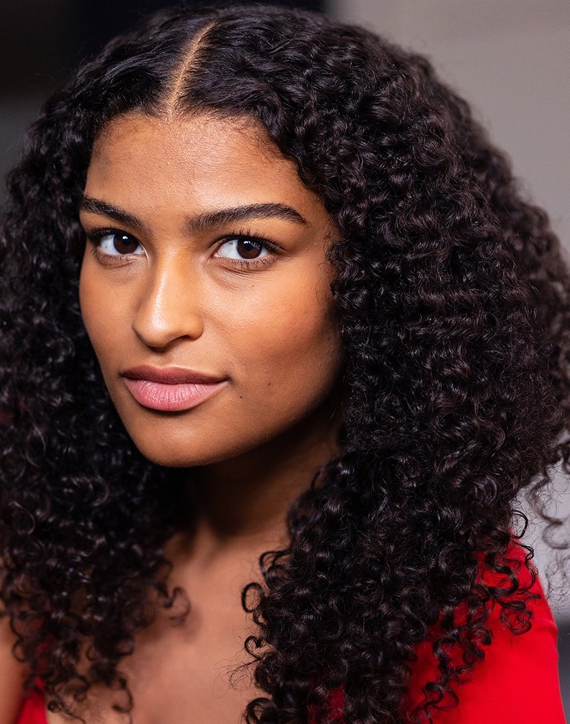 Close-up portrait of a young woman with dark, curly hair and brown eyes, wearing a red top, looking directly at the camera with a slight smile.