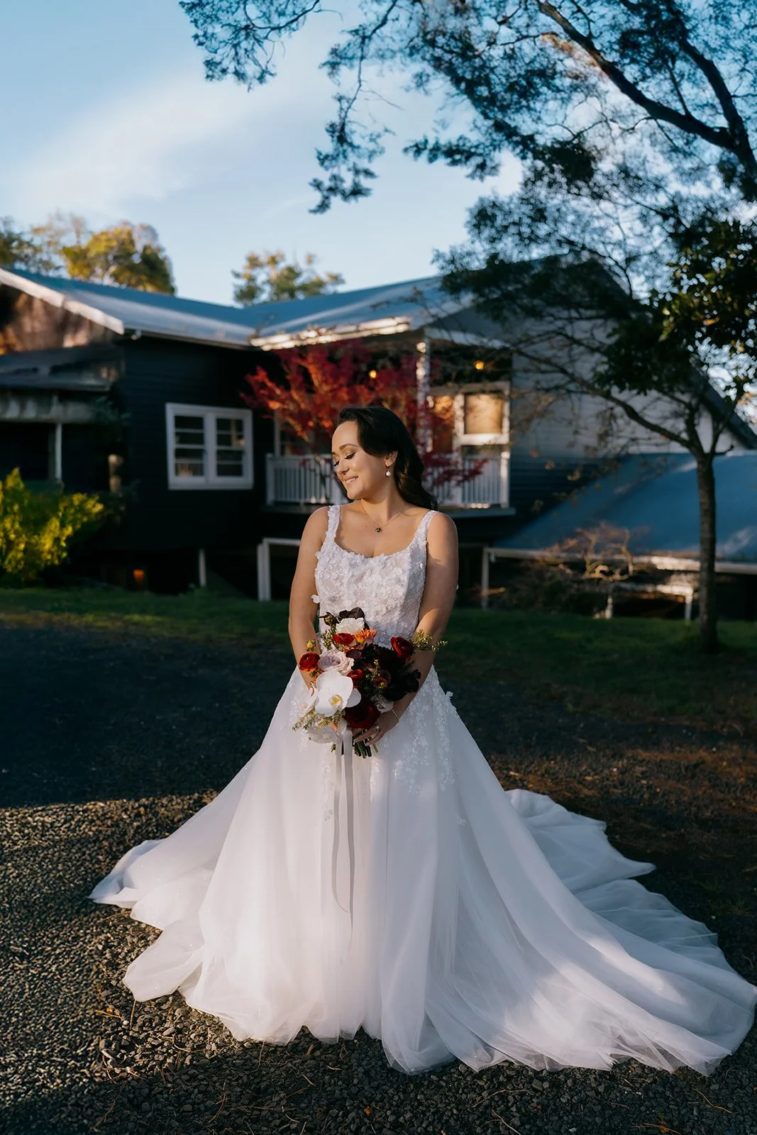 A bride in a white wedding gown holding a bouquet, standing outside during sunset in front of a house with trees.