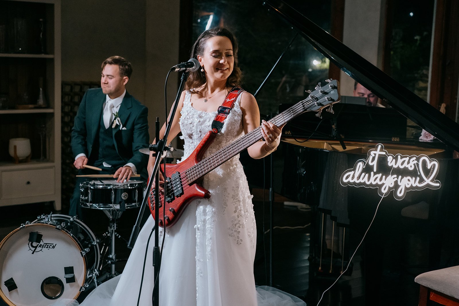 A woman in a white wedding dress playing an electric guitar, with a man in a suit on drums behind her, and a pianist at a grand piano with a neon sign that reads 'trust always you' in the background.