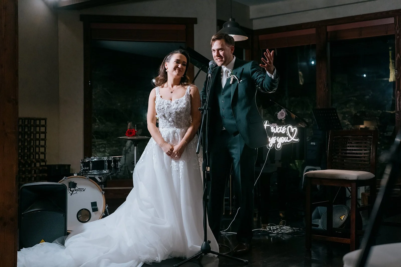 A bride and groom giving a speech at their wedding reception, standing in front of a microphone with musical instruments and decorative lights in the background.