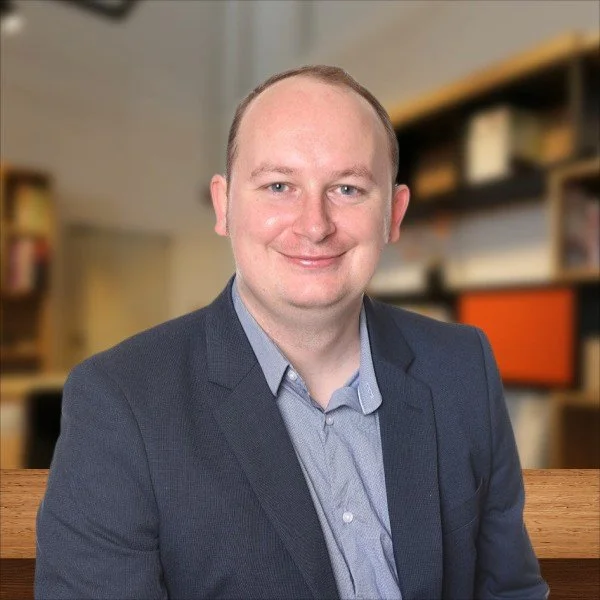 A man with short light brown hair and fair skin wearing a dark blazer and a light blue collared shirt, sitting in a room with blurred bookshelves in the background.