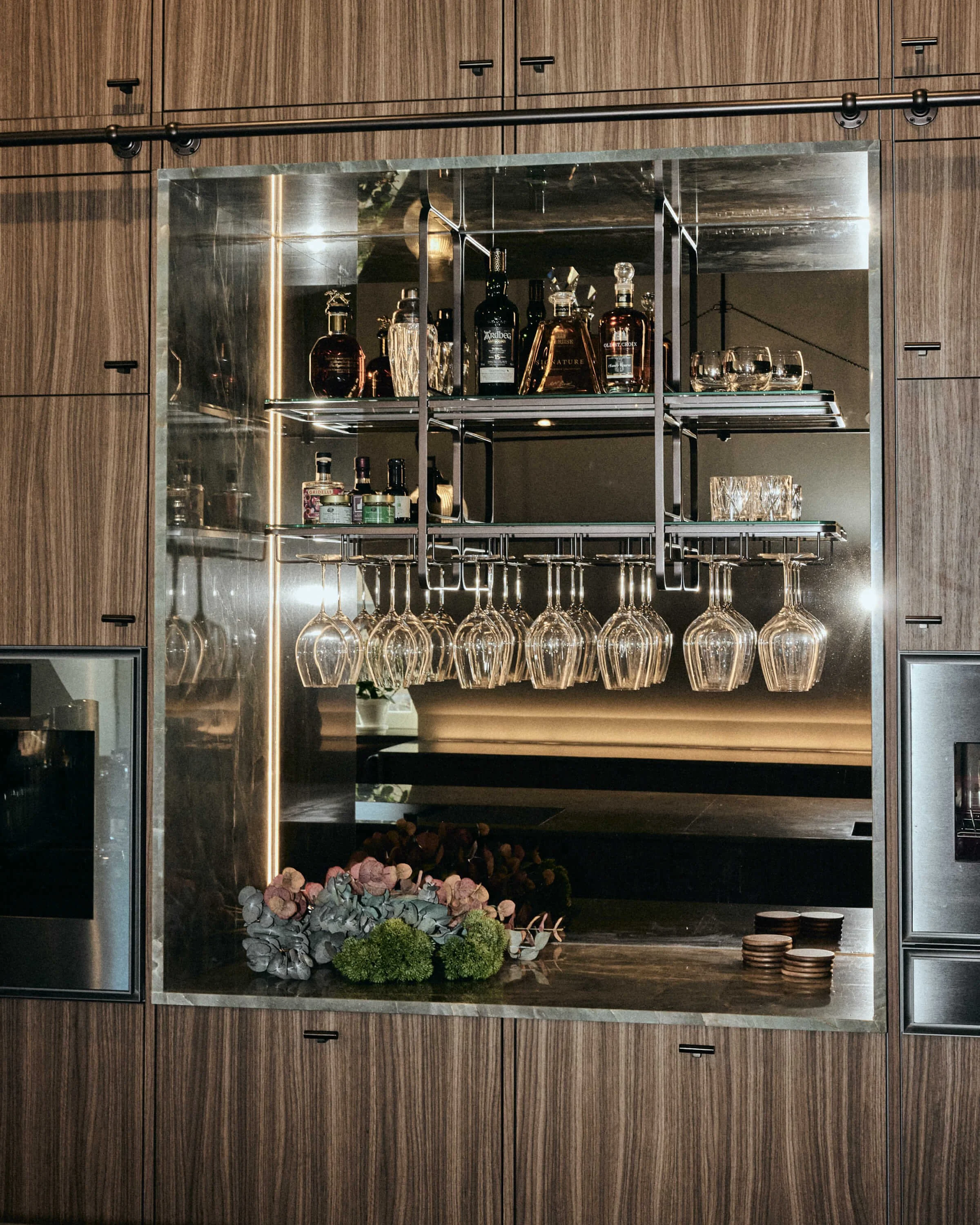 A built-in bar cabinet with wooden panels, mirrored interior, wine glasses hanging upside down, bottles of liquor on shelves, and decorative items including flowers and coasters.