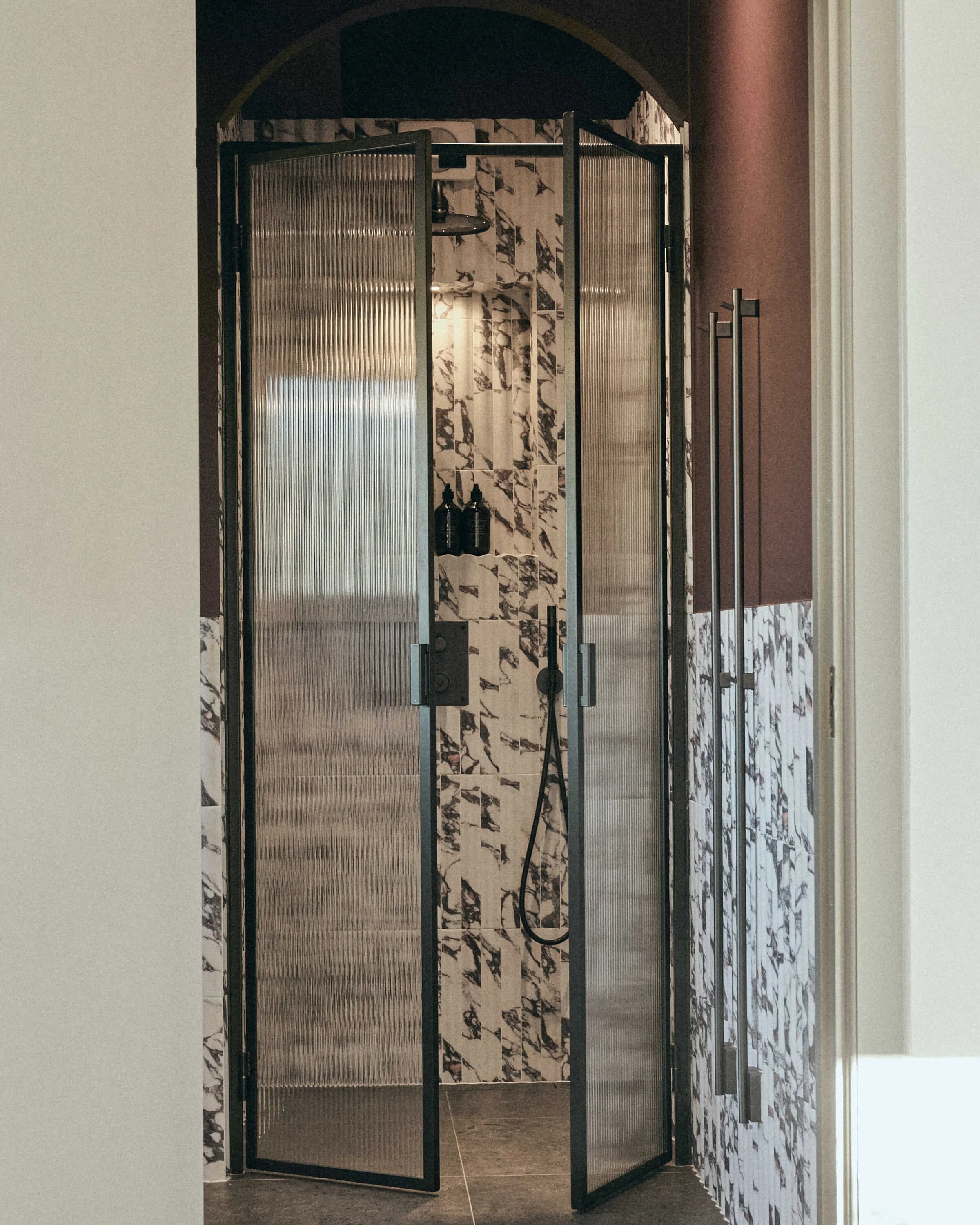 Modern shower with black framed glass door, black wall-mounted showerhead, and black bottles on marble and tile wall.