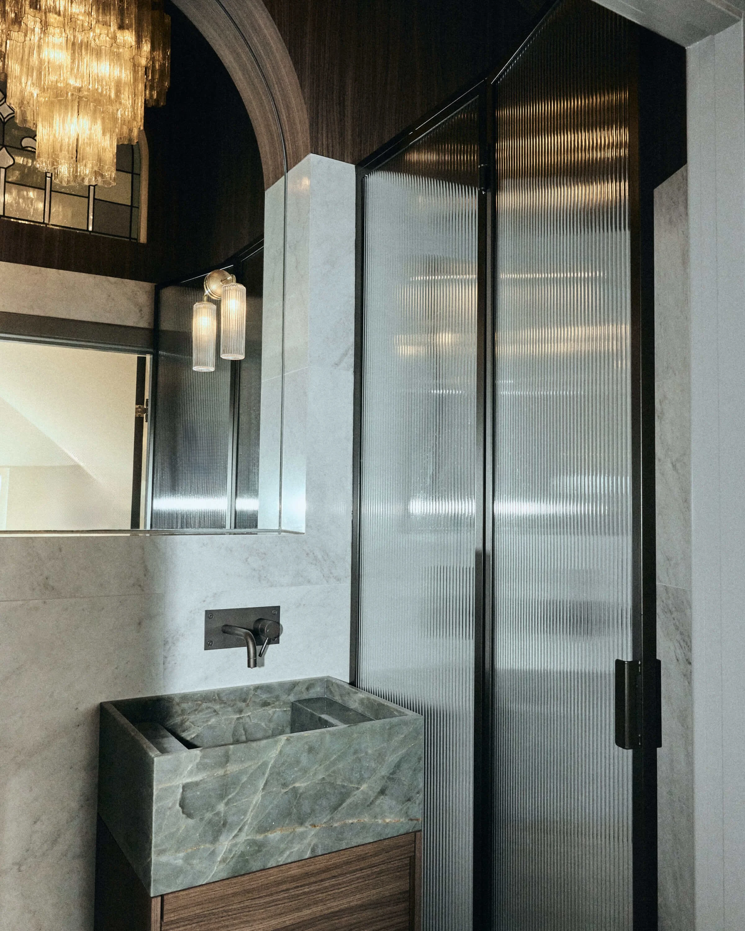 Modern bathroom with marble sink, wall-mounted black faucet, and ribbed metallic sliding door. Elegant lighting fixtures hang from the ceiling.