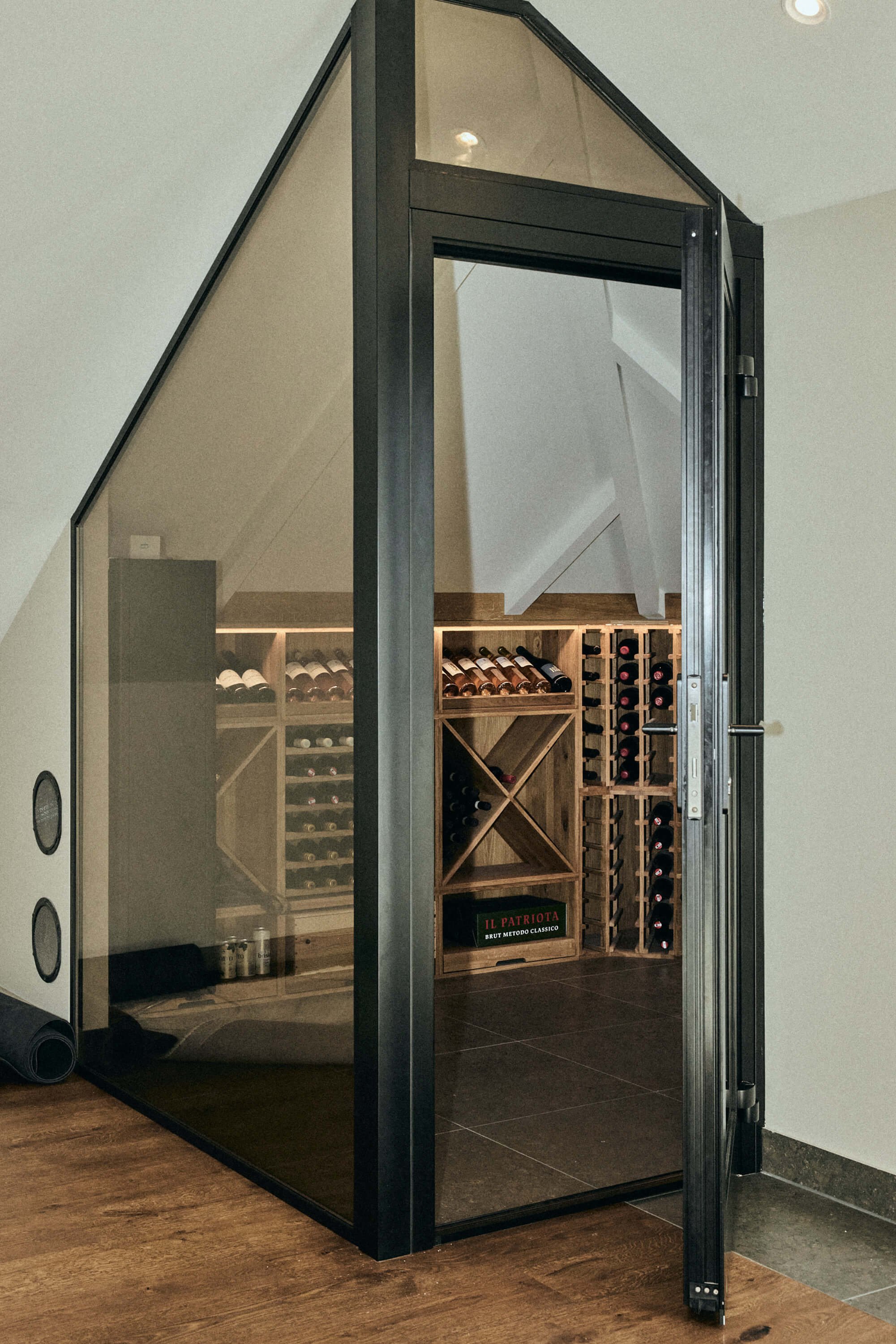 Glass wine cellar with wooden shelves filled with wine bottles, located in an attic space with sloped ceiling.