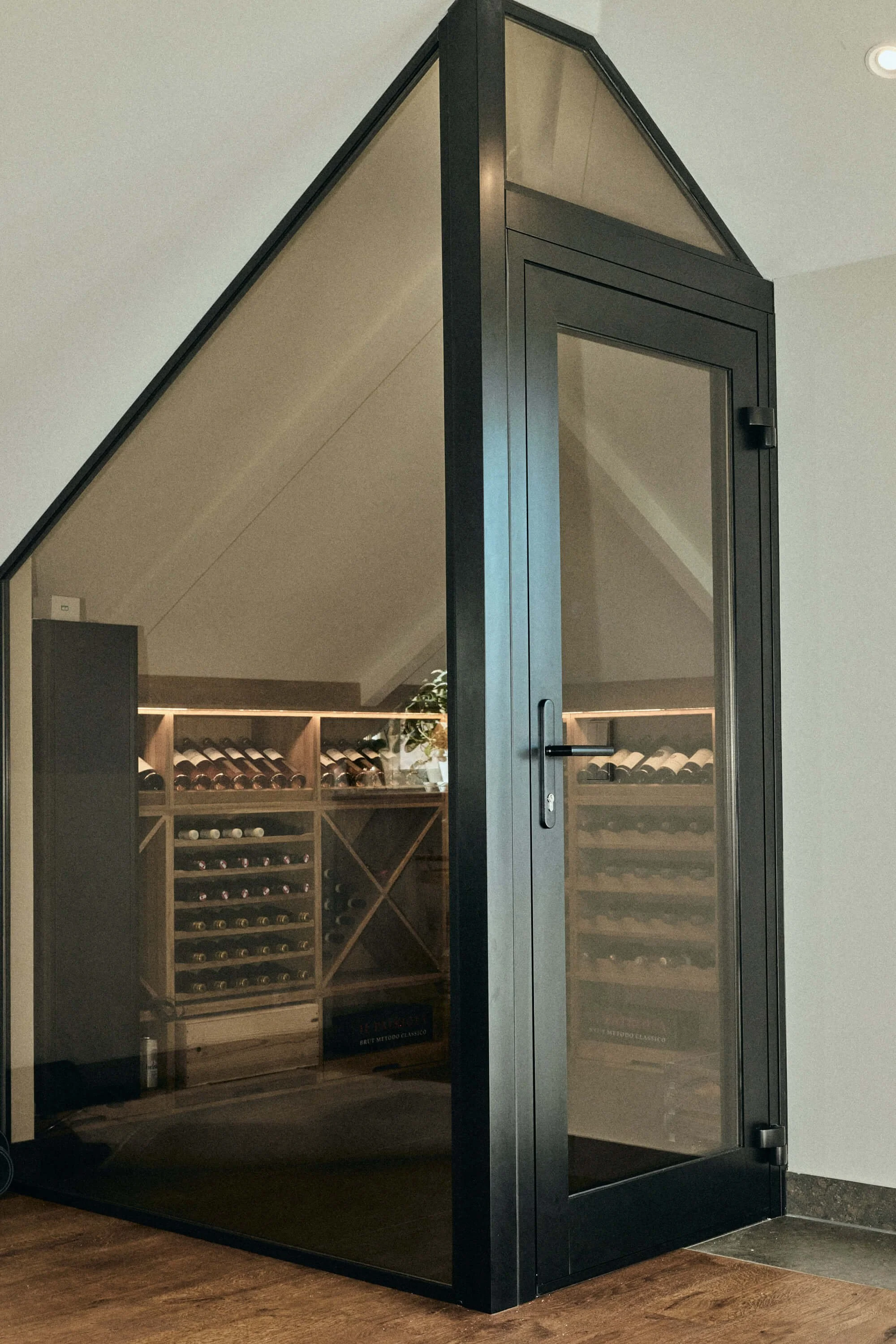 Glass wine cellar with wooden wine racks and bottles inside, located in a room with a sloped ceiling.