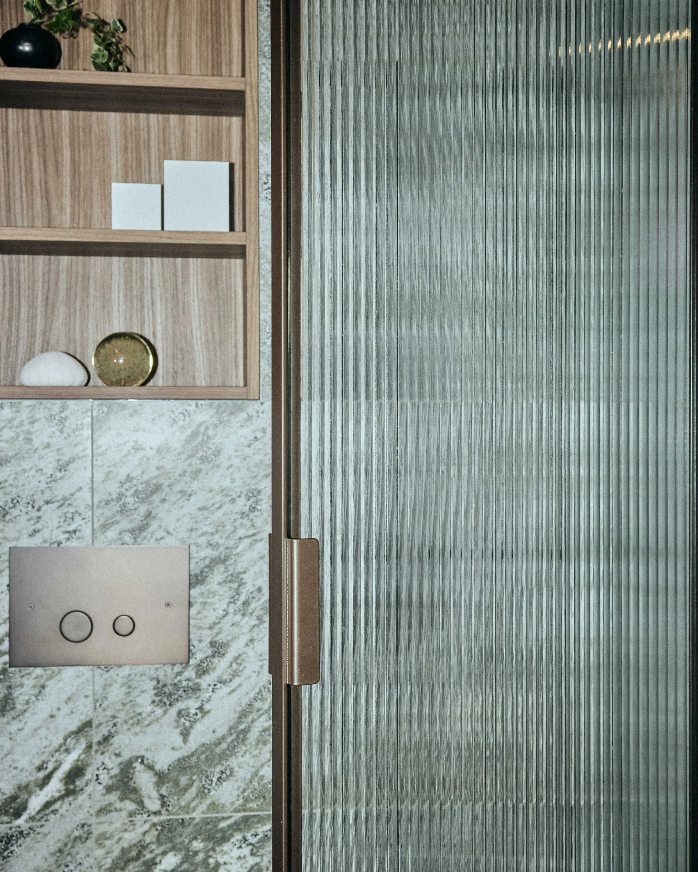 Part of a bathroom with a frosted glass shower door, a wooden shelving unit with decorative items, and a marble wall with a toilet flush button.