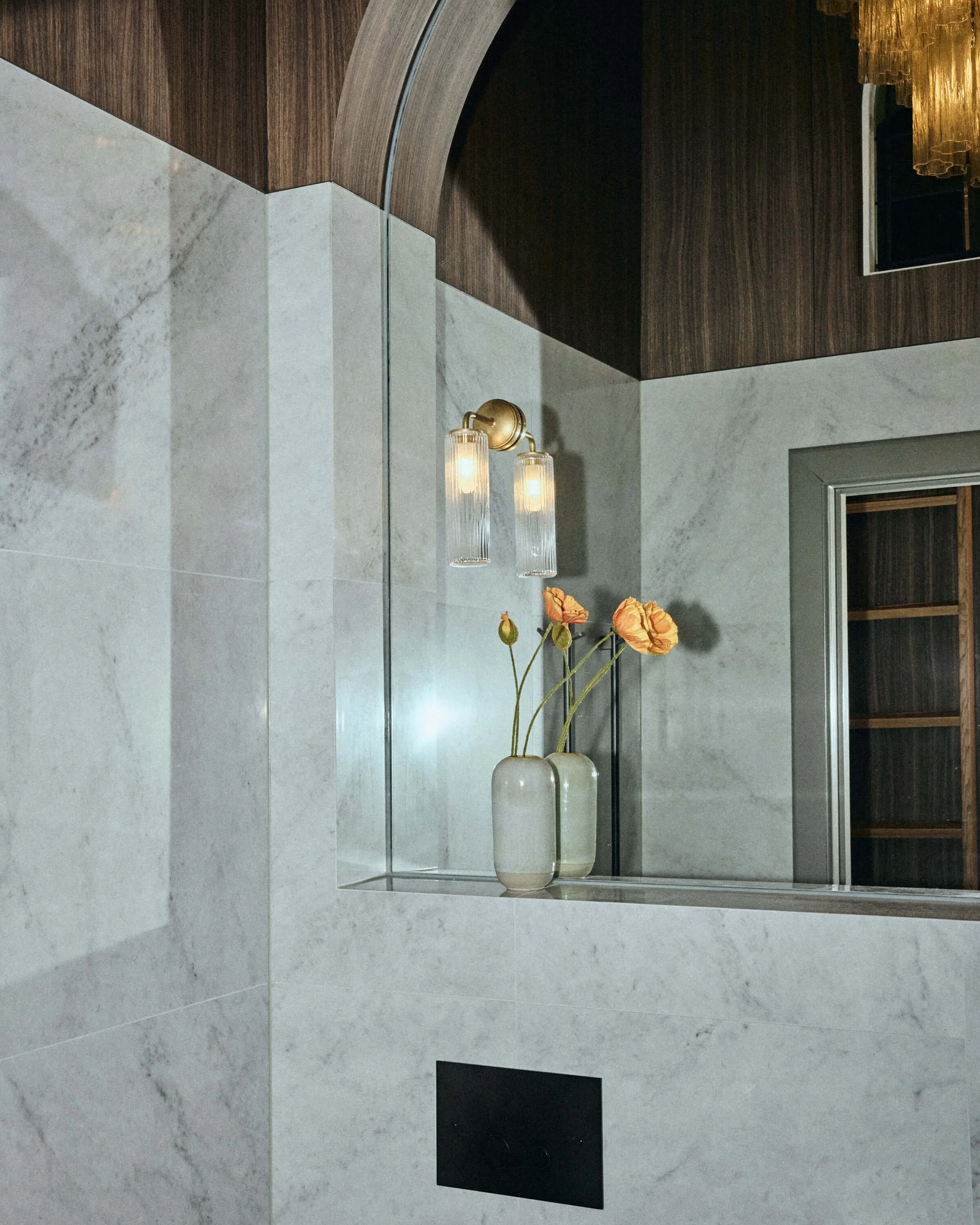 Interior wall with marble panel, decorative vases with orange flowers, wooden and metal window frame, glass wall sconce lights, and a mirror reflecting a chandelier.