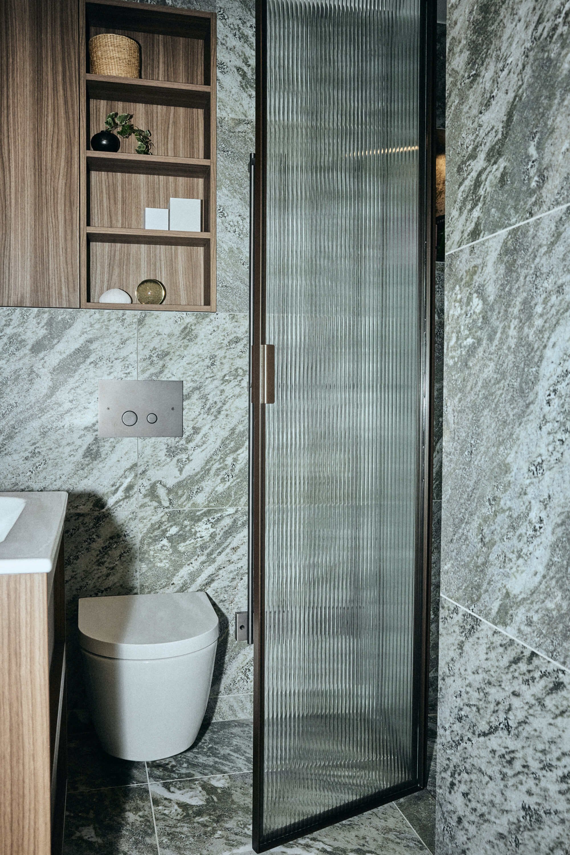 Modern bathroom with a frosted glass door, wall-mounted toilet, and wooden shelving with decorative items.