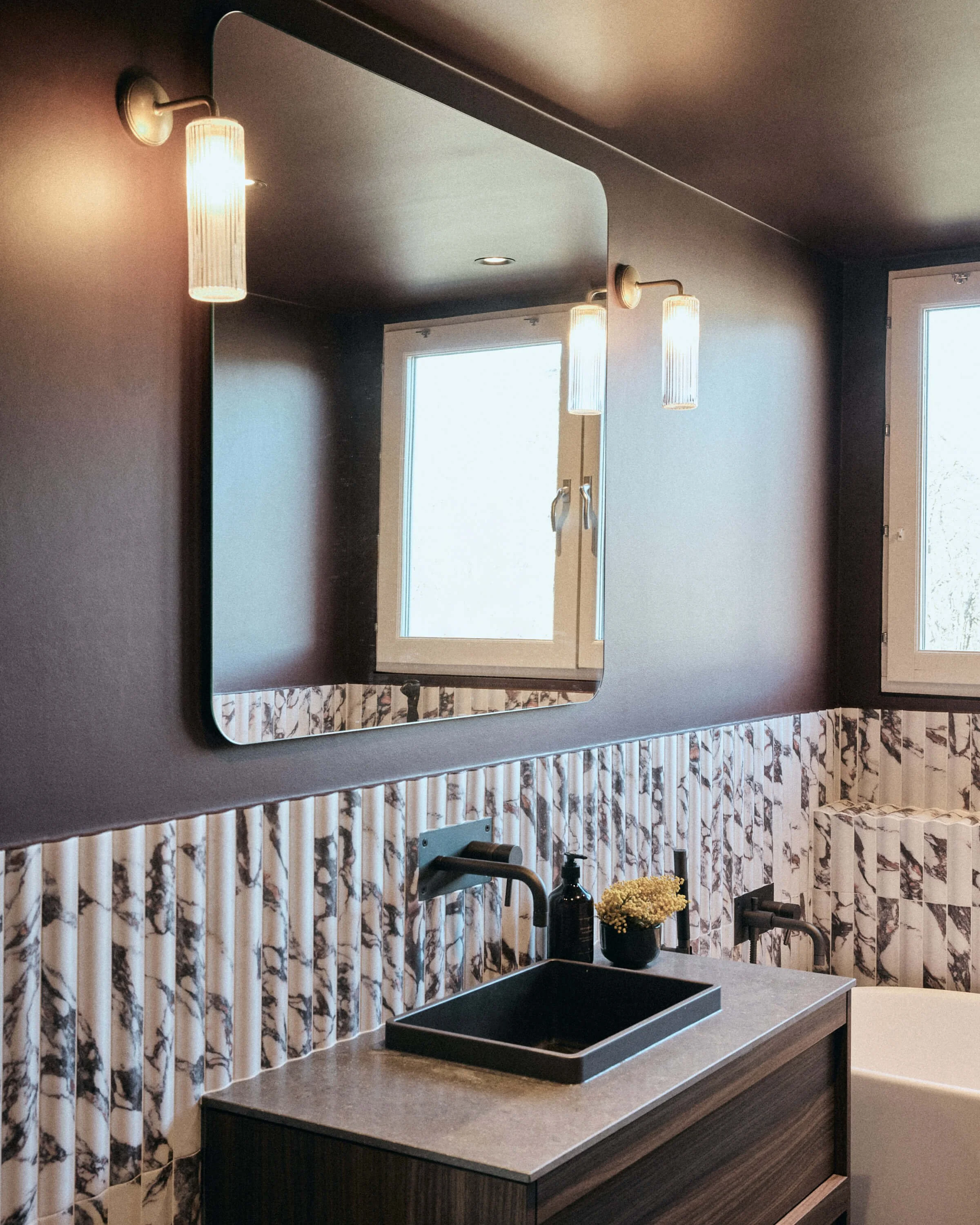 Bathroom with a black sink, a mirror, wall-mounted lamps, a window, and marble-patterned wainscoting.