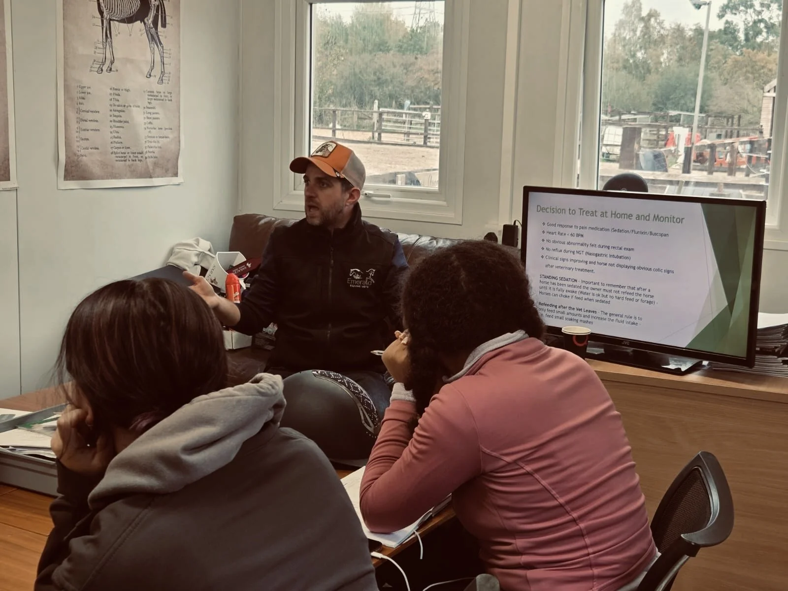 A man giving a presentation about horse treatment to a group of three women in a room with large windows and a television screen displaying a slide titled 'Decision to Treat at Home and Monitor'.