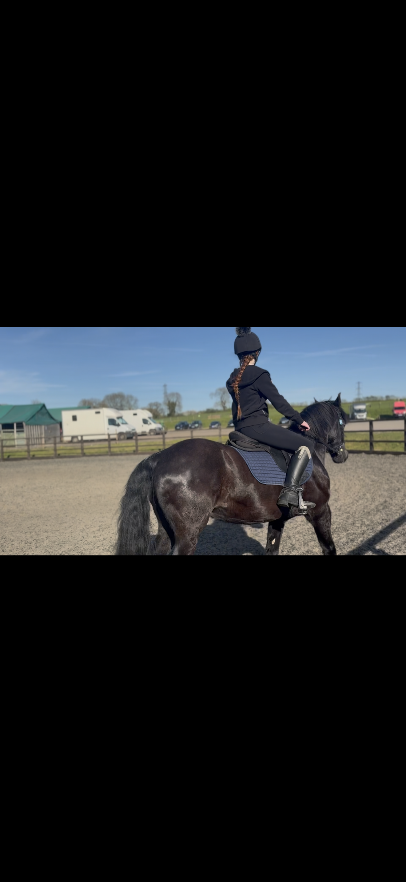 A young girl riding a black horse in an outdoor riding arena on a sunny day, with trailers and parked cars visible in the background.