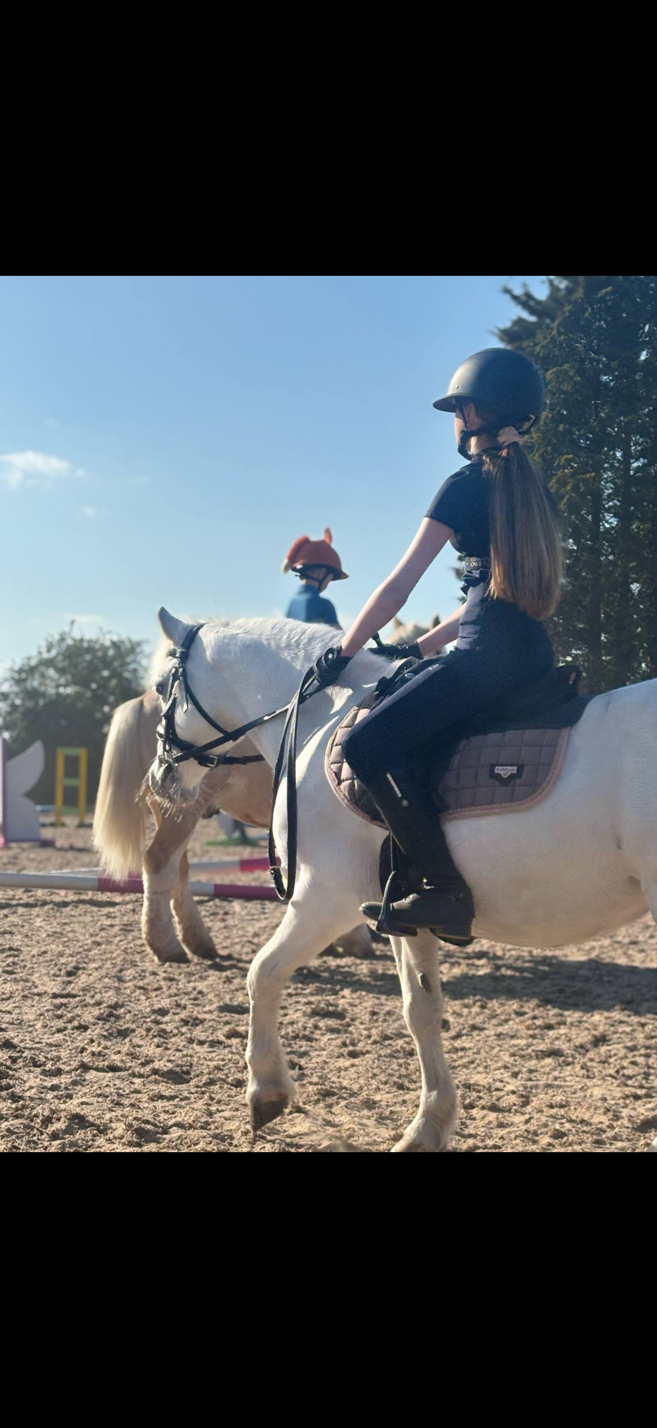 Young girl riding a white horse in an outdoor riding arena, wearing a helmet and riding gear, under a clear blue sky.