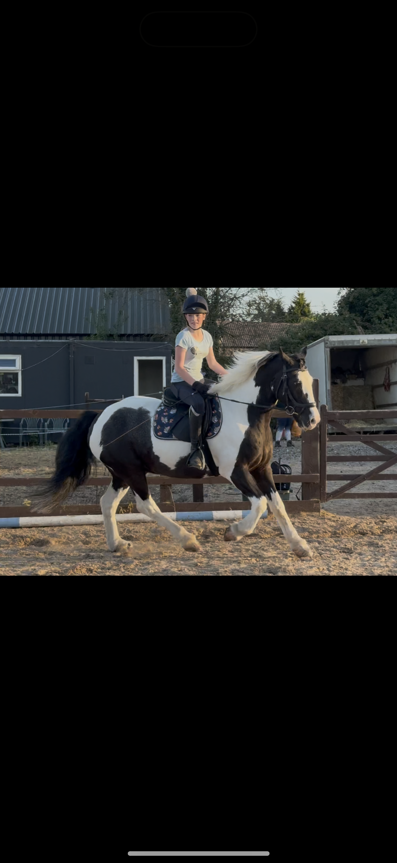 A young girl wearing a helmet riding a black and white pinto horse in an outdoor riding arena during sunset.