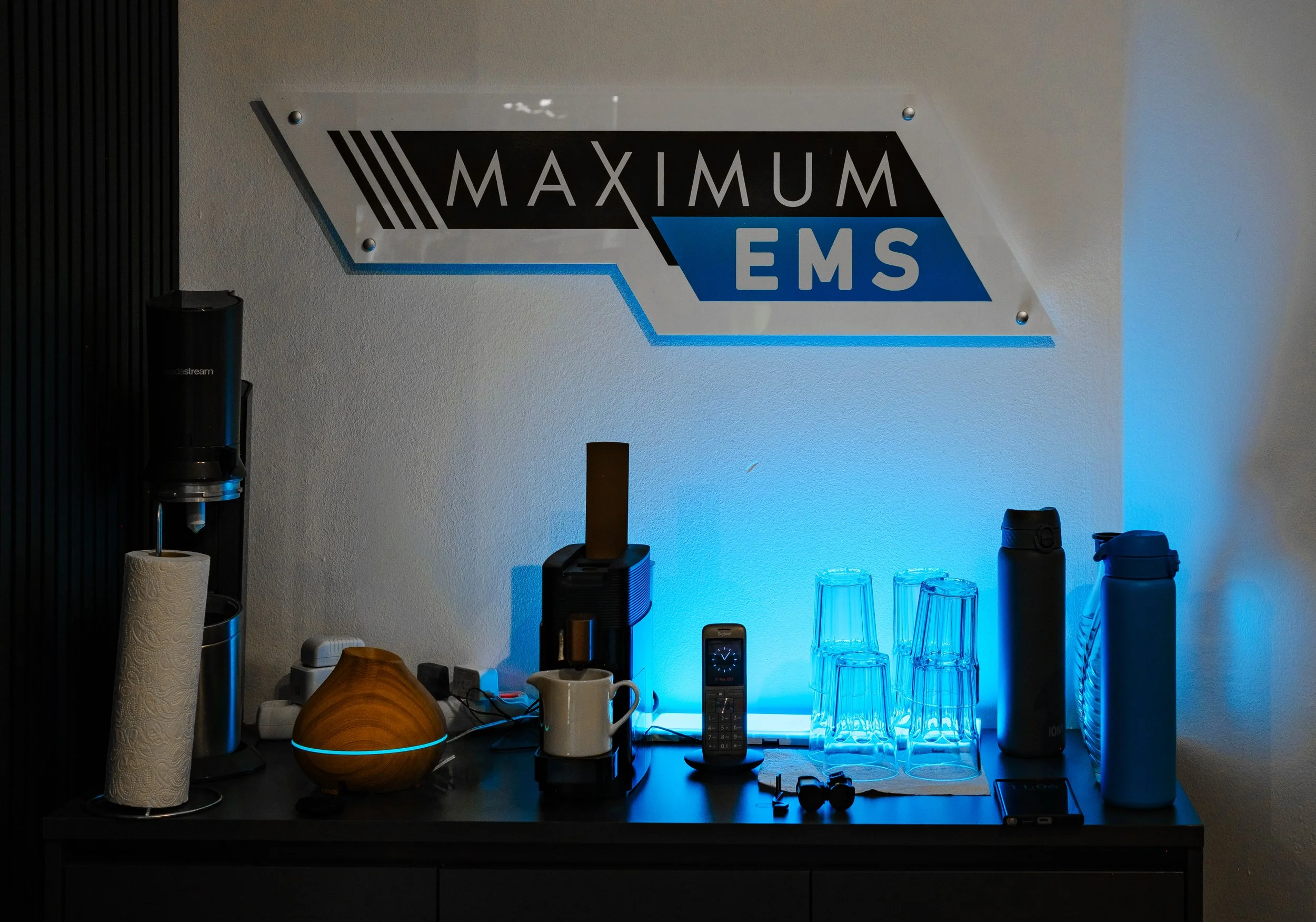 Getränkestation mit Wasserflaschen, Gläsern, Kaffeemaschine, Wasserkocher, Lautsprecher, Handy und anderen Gegenständen, vor einer Wand mit einer Maximum EMS Beschilderung.