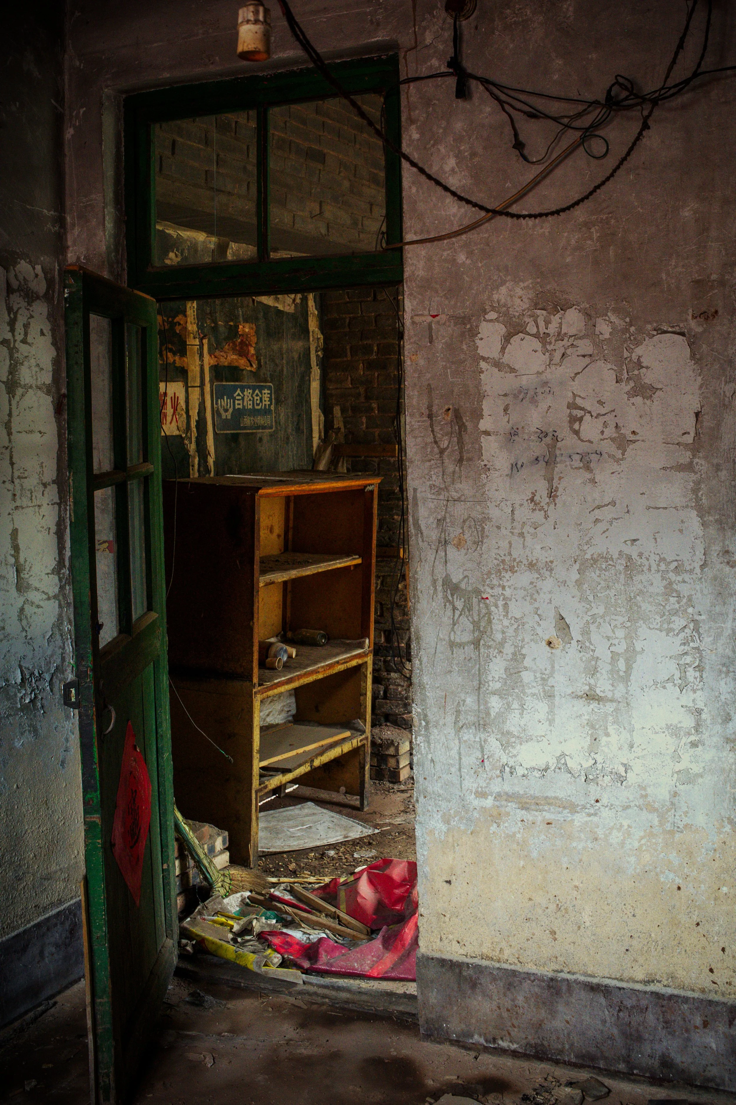 An abandoned room with peeling paint on the walls, scattered debris on the floor, a cluttered wooden shelf, and a window with green trim revealing brick architecture outside. Electrical wires are exposed along the ceiling.
