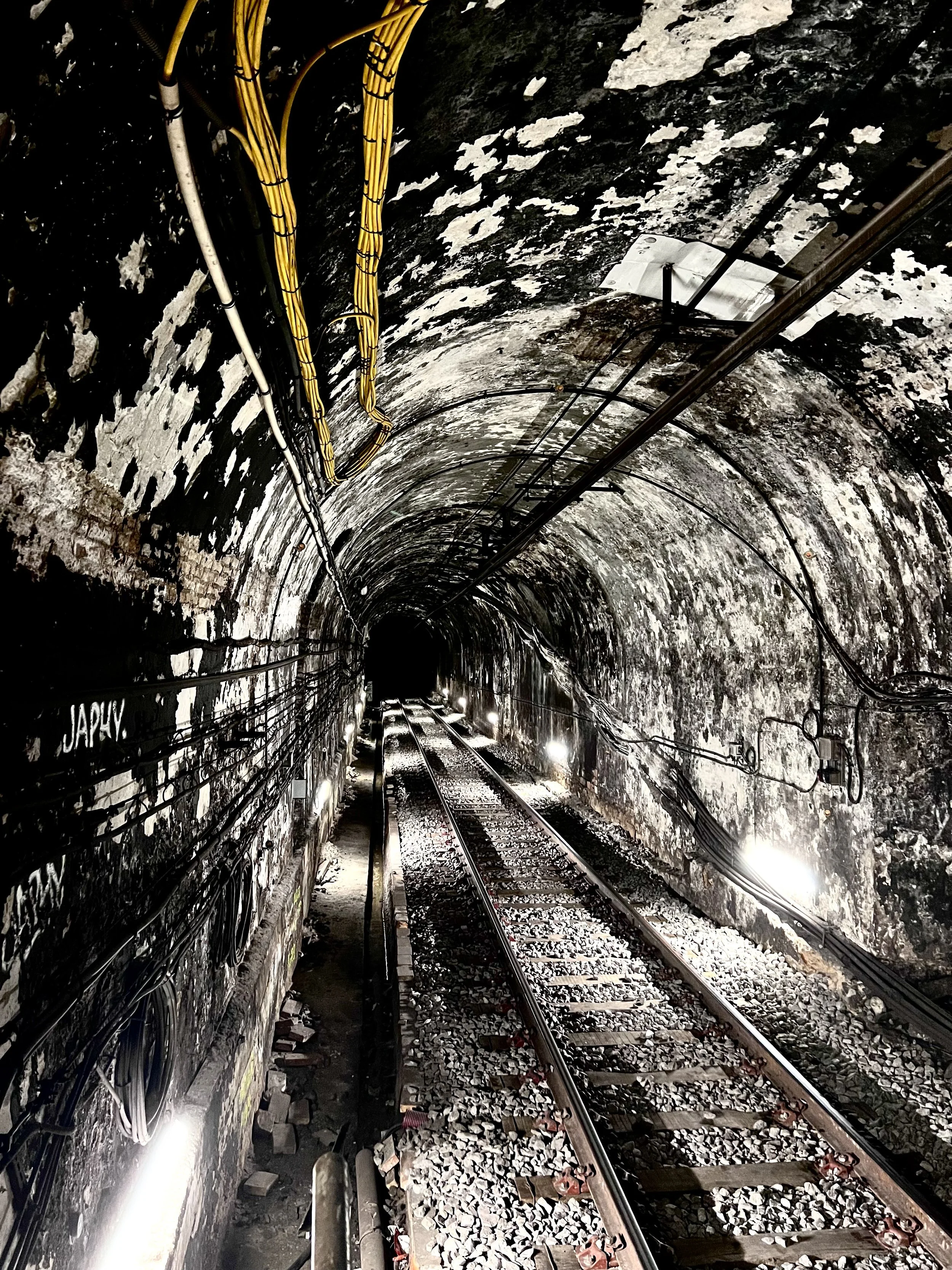 Perth street photography. Inside a tunnel with railway tracks and electrical wiring along the rough, dark, and weathered rock walls, illuminated by small lights.