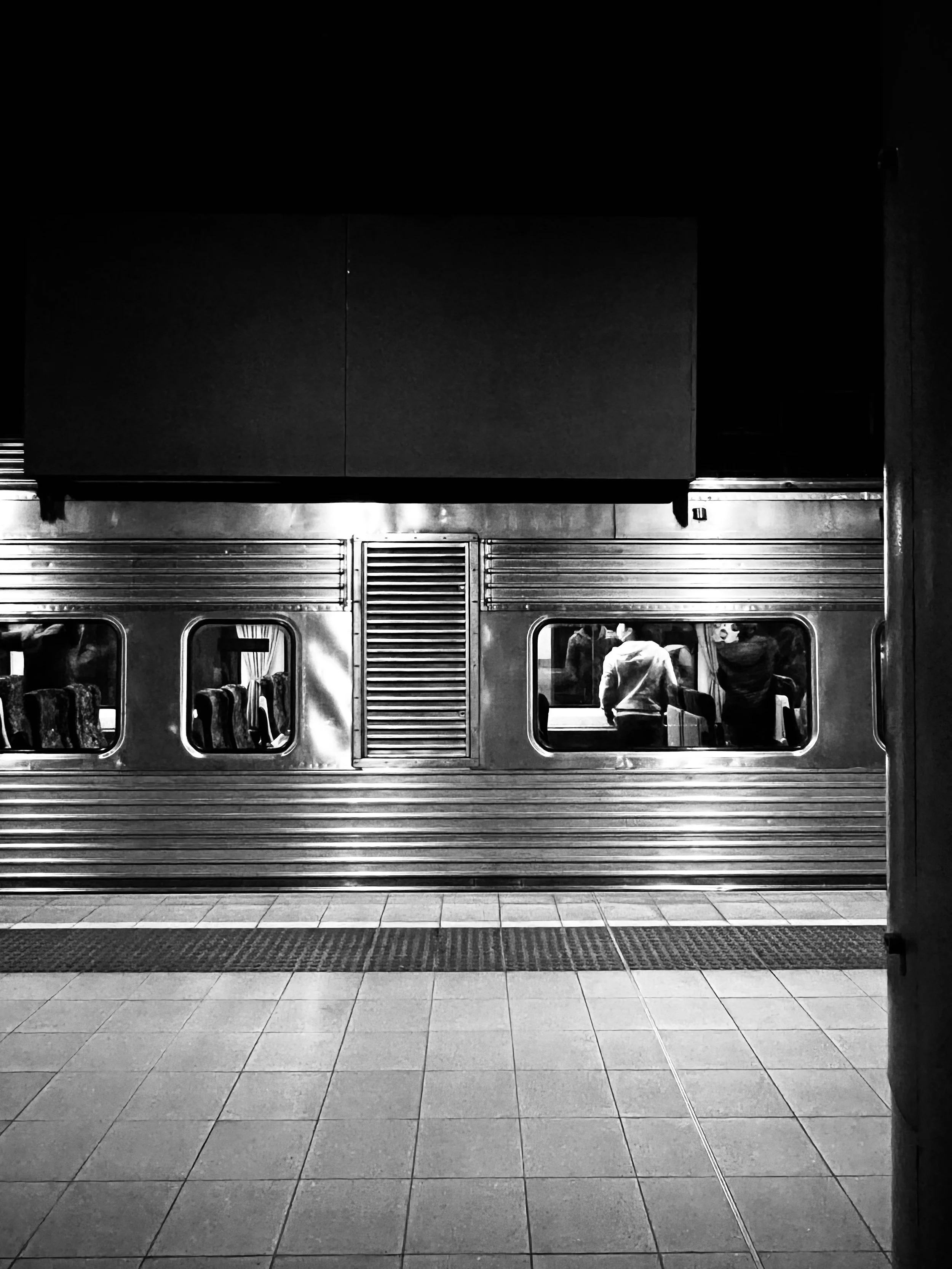 Perth street photography Black and white photo of a train station platform at night with a reflective metal train, showing two people inside the train through the windows. The platform is tiled with a textured strip for safety.