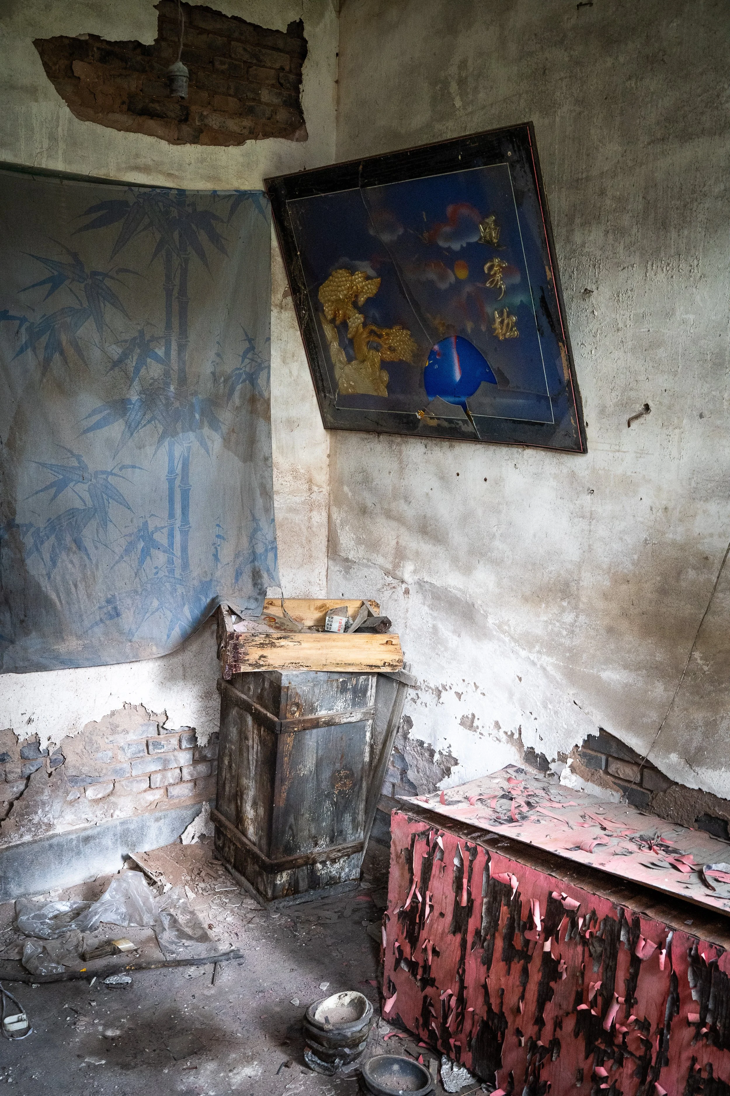 An abandoned room with peeling plaster walls, a broken desk, and debris on the dusty floor. There's a large, shattered painting with Chinese characters on the wall, a wooden box and several small empty bowls or pots on the floor.