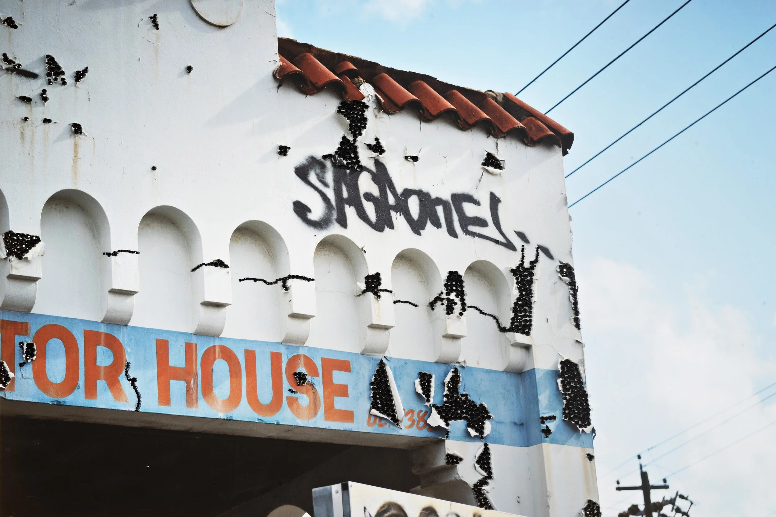 Perth Kintsugi Exhibition- Photograph with beads A building with graffiti and numerous black clusters of insects attached to its white walls, with part of a blue and orange sign reading '& for house' and a red-tiled roof, under a partly cloudy sky.