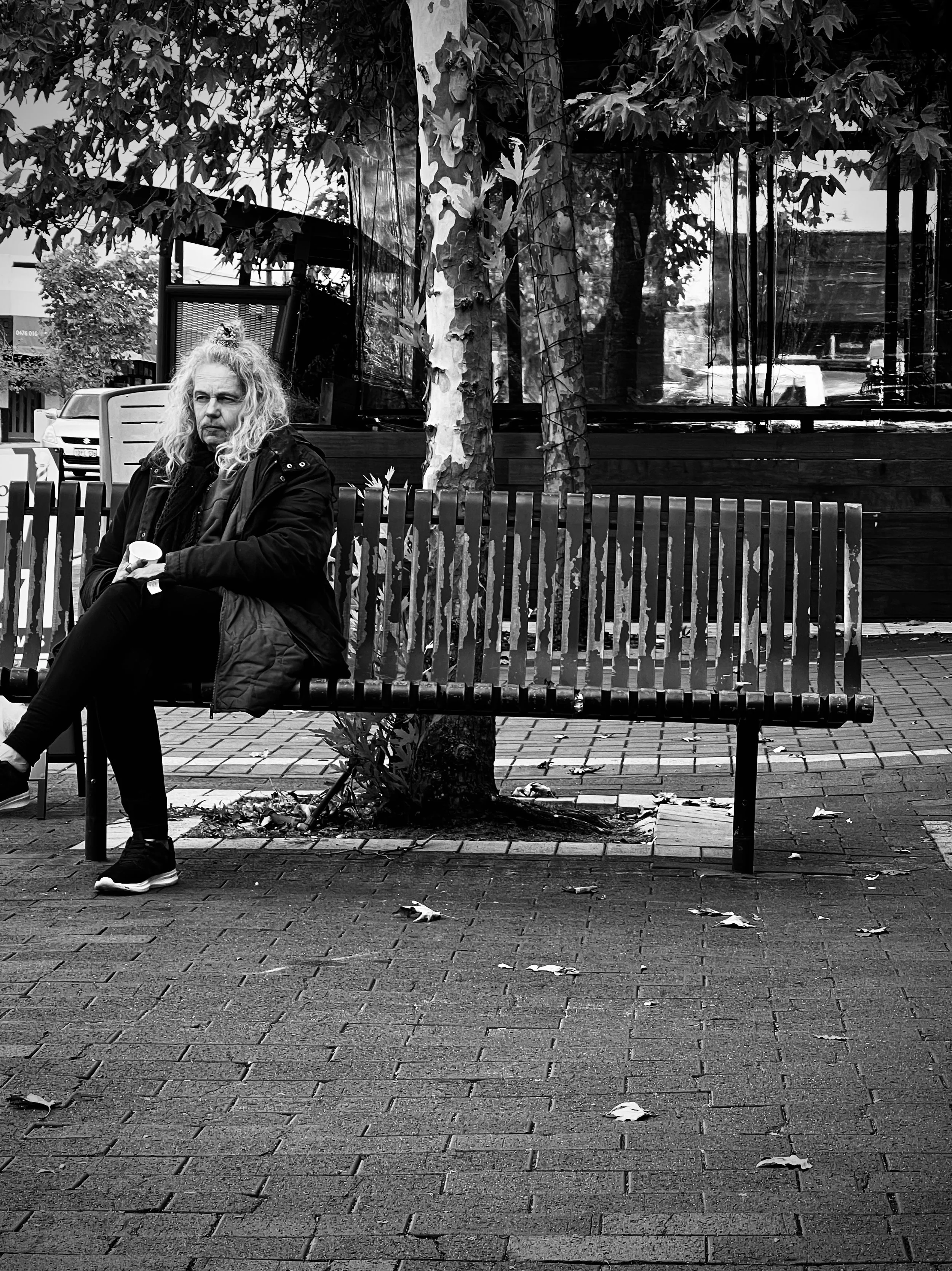 Perth street photography. A woman with long curly hair sitting alone on a park bench holding a cup, wearing dark clothing. There are trees and a building in the background, and fallen leaves on the ground.
