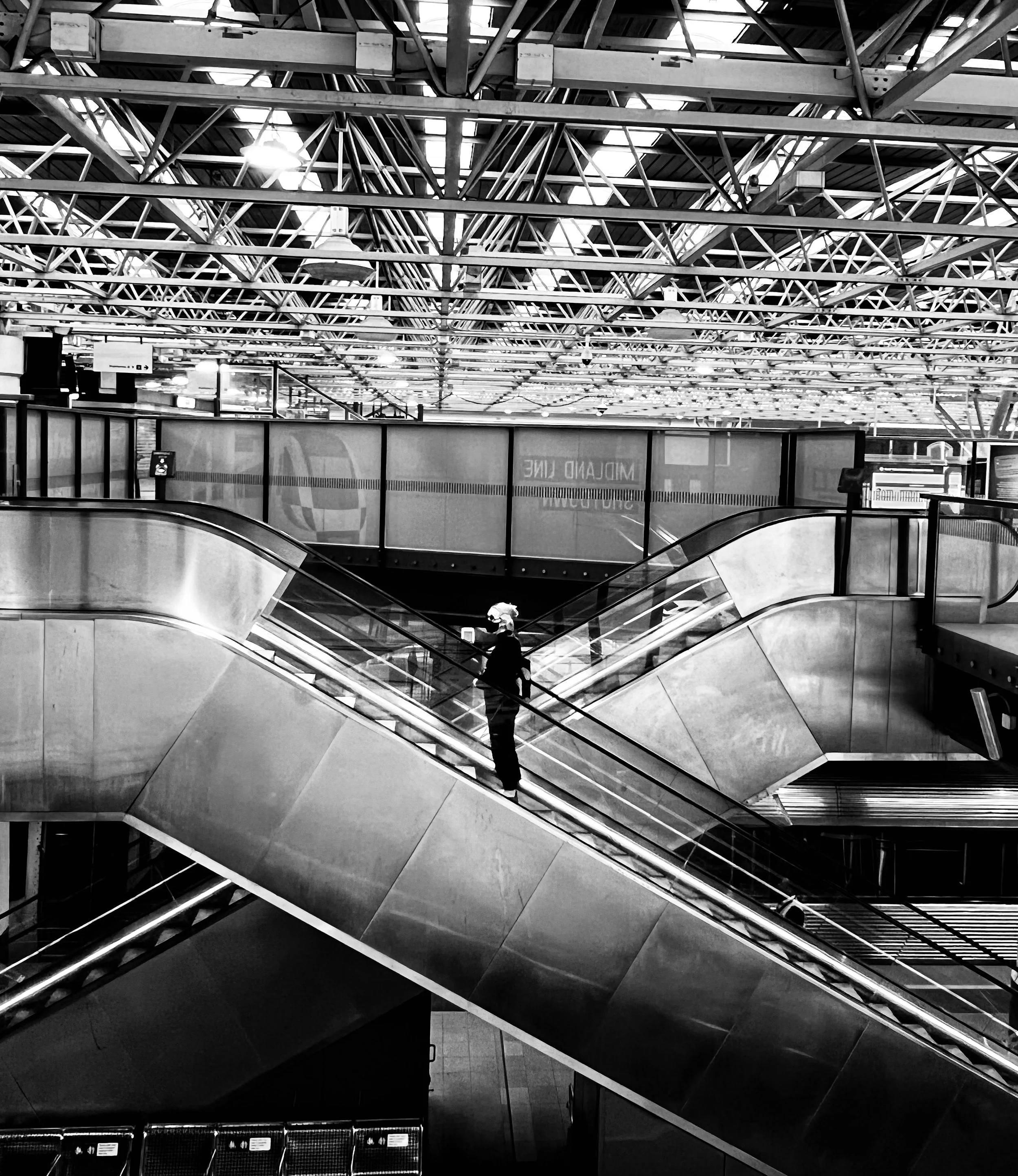 Perth street photography A person standing on a moving walkway in a modern, industrial-style transportation terminal with a high ceiling and exposed metal trusses, captured in black and white.