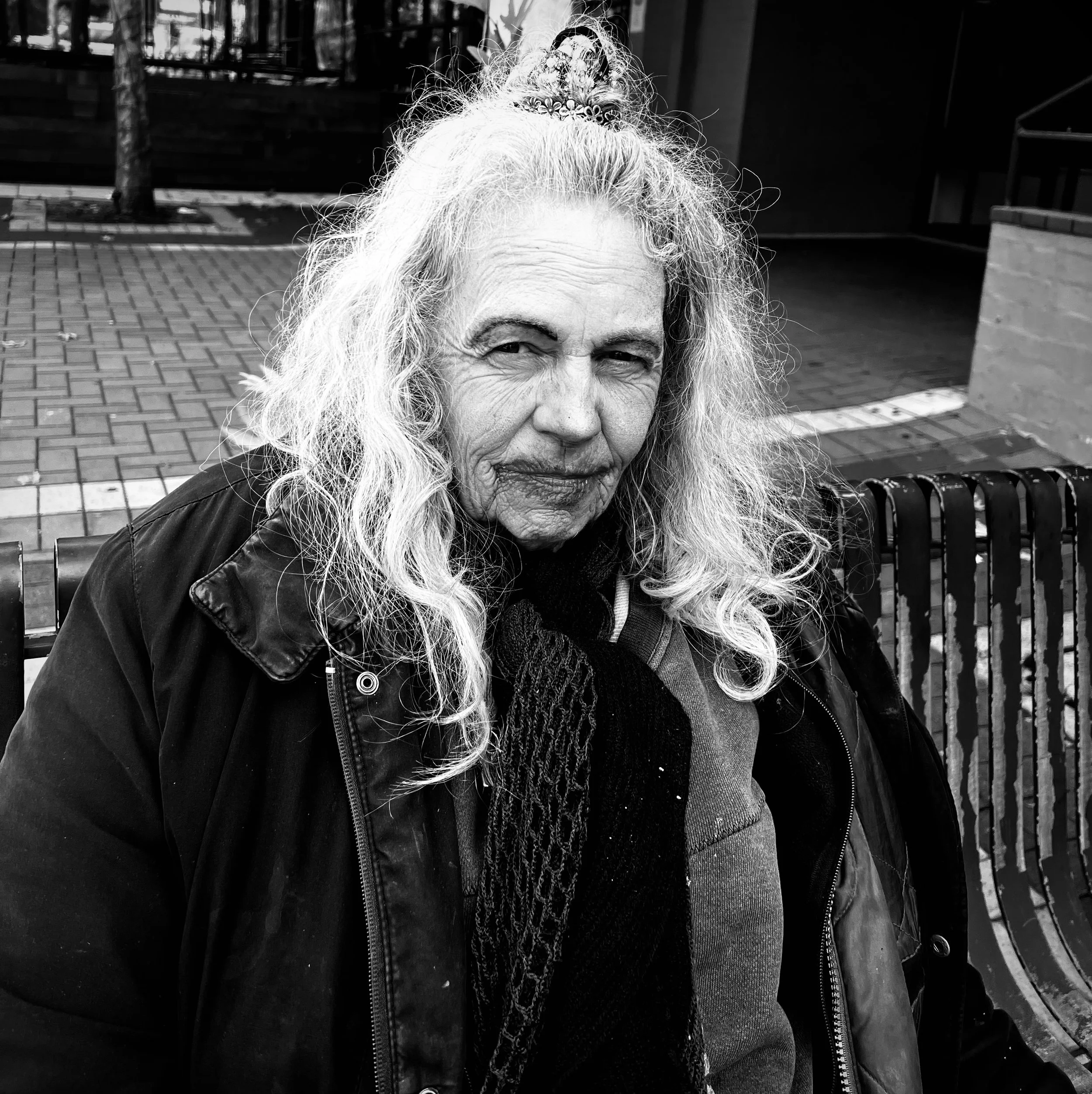 Perth street photography-Black and white photograph of an elderly woman with long curly hair sitting on a park bench. She has a slightly mischievous expression, wearing a jacket and scarf, with a background of pavement and a brick wall.