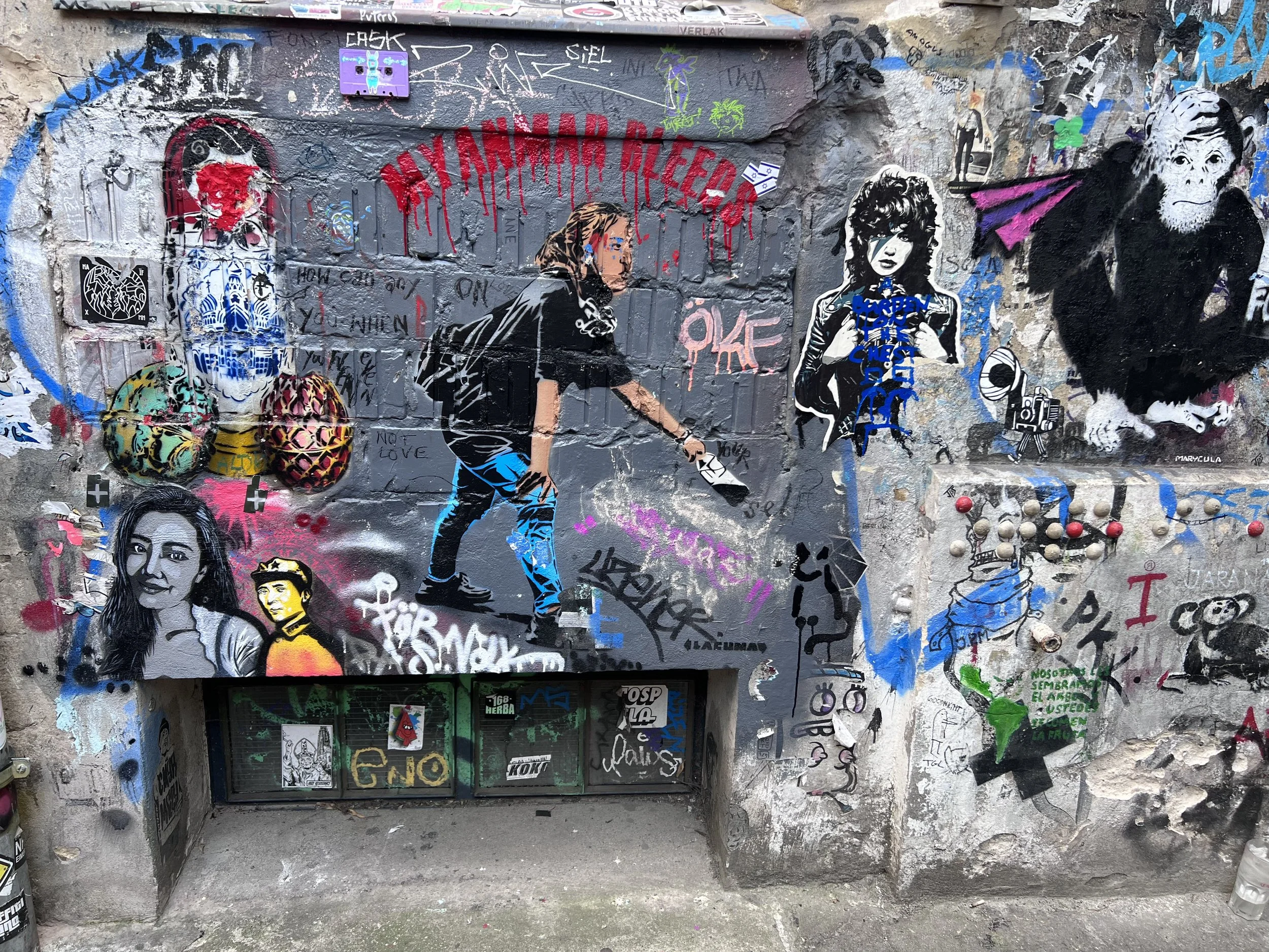 Berlin Street- Street art graffiti wall with various colorful images, including a girl spray painting on wall, stylized portraits of a woman and an ape, graffiti tags, and stickers.