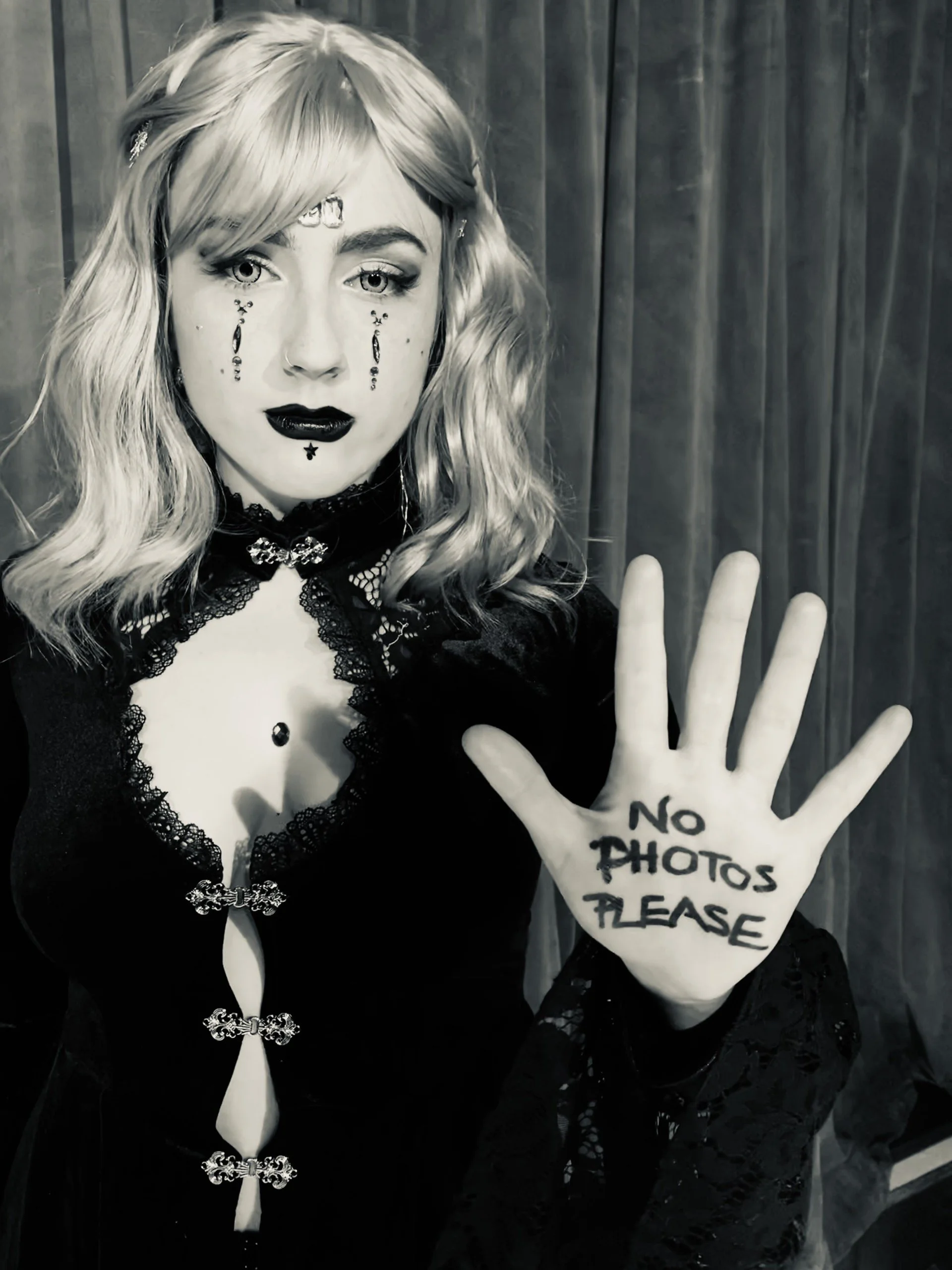 A person with wavy blonde hair and gothic makeup holding up their hand with the words 'NO PHOTOS PLEASE' written on it. The person is wearing a black lace and satin outfit with decorative buttons, dark lipstick, and makeup. The background is a curtai