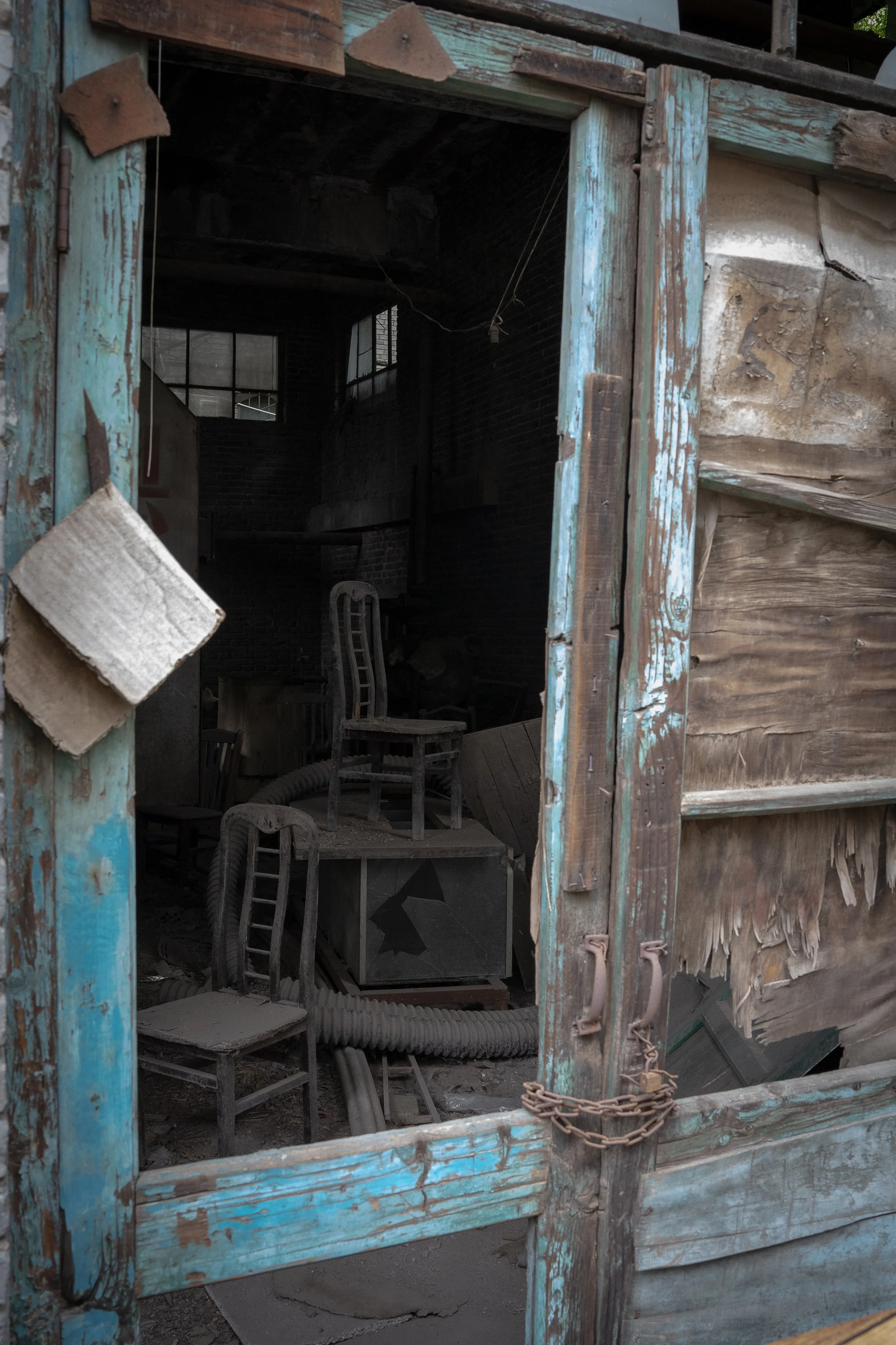 An old, weathered wooden door with peeling blue paint, secured with a rusty chain, opens to an abandoned space filled with dust, old chairs, and debris. The interior appears dark with brick walls and a window visible in the background.