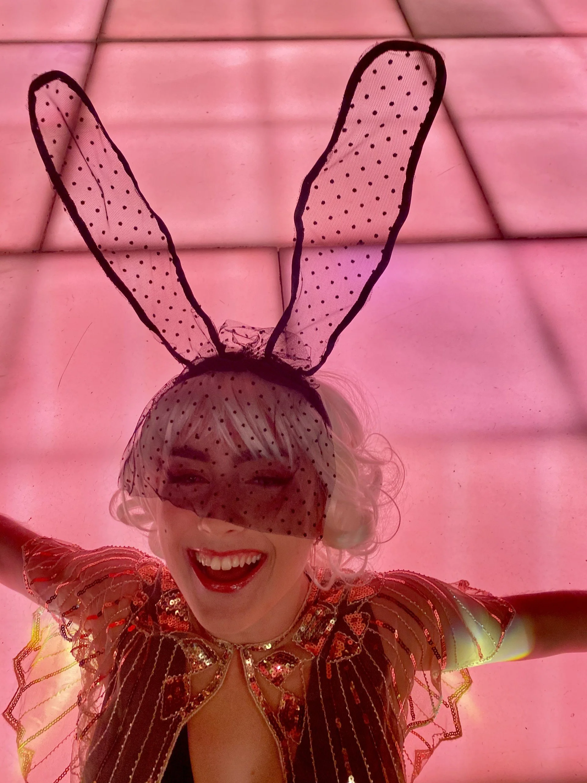 A woman with curly blonde hair wearing bunny ears and a polka dot veil, smiling and wearing a colorful, sequined outfit with a butterfly design.