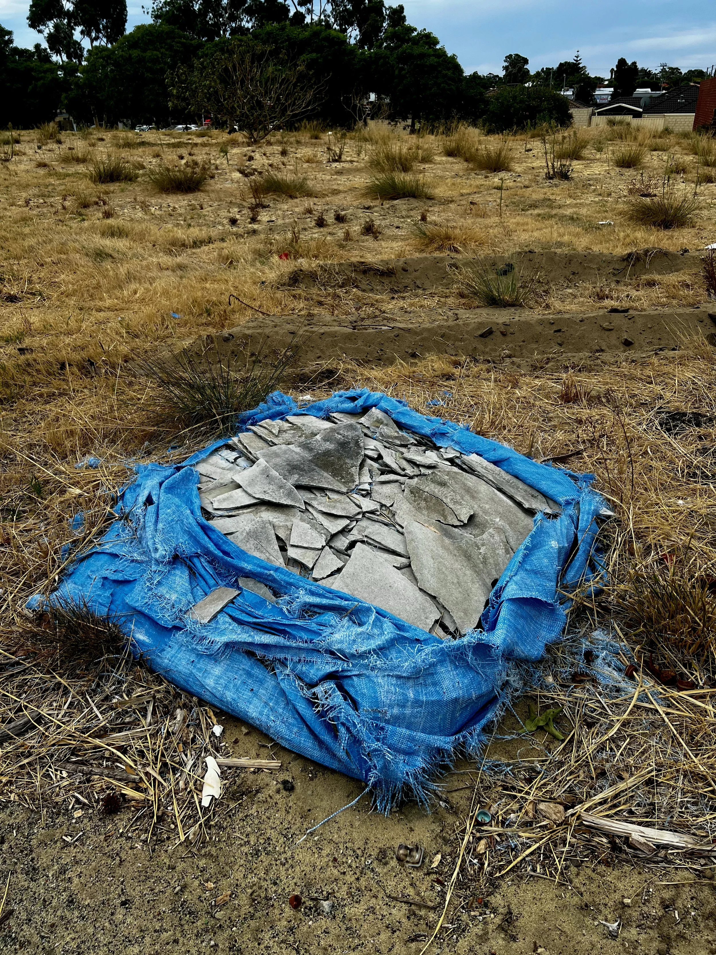 Perth street photography- A pile of asbestos on a blue tarp, placed on dry grass with sparse vegetation in an open field. 