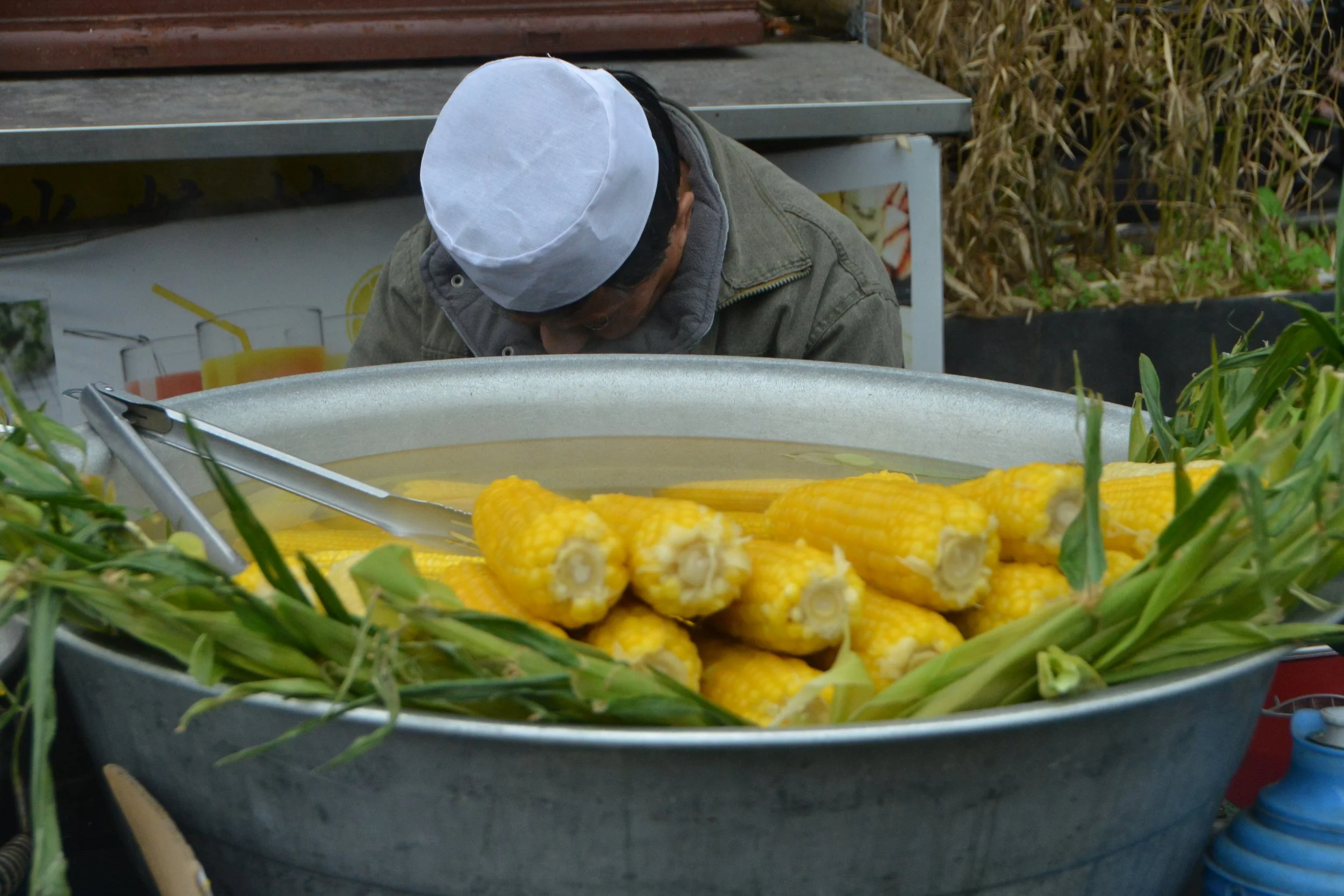 Part of the China Exhibition-Street Photography. A man wearing a white hat and gray jacket cooking corn on the cob in a large pot outdoors, with some green leaves and corn husks around the pot.