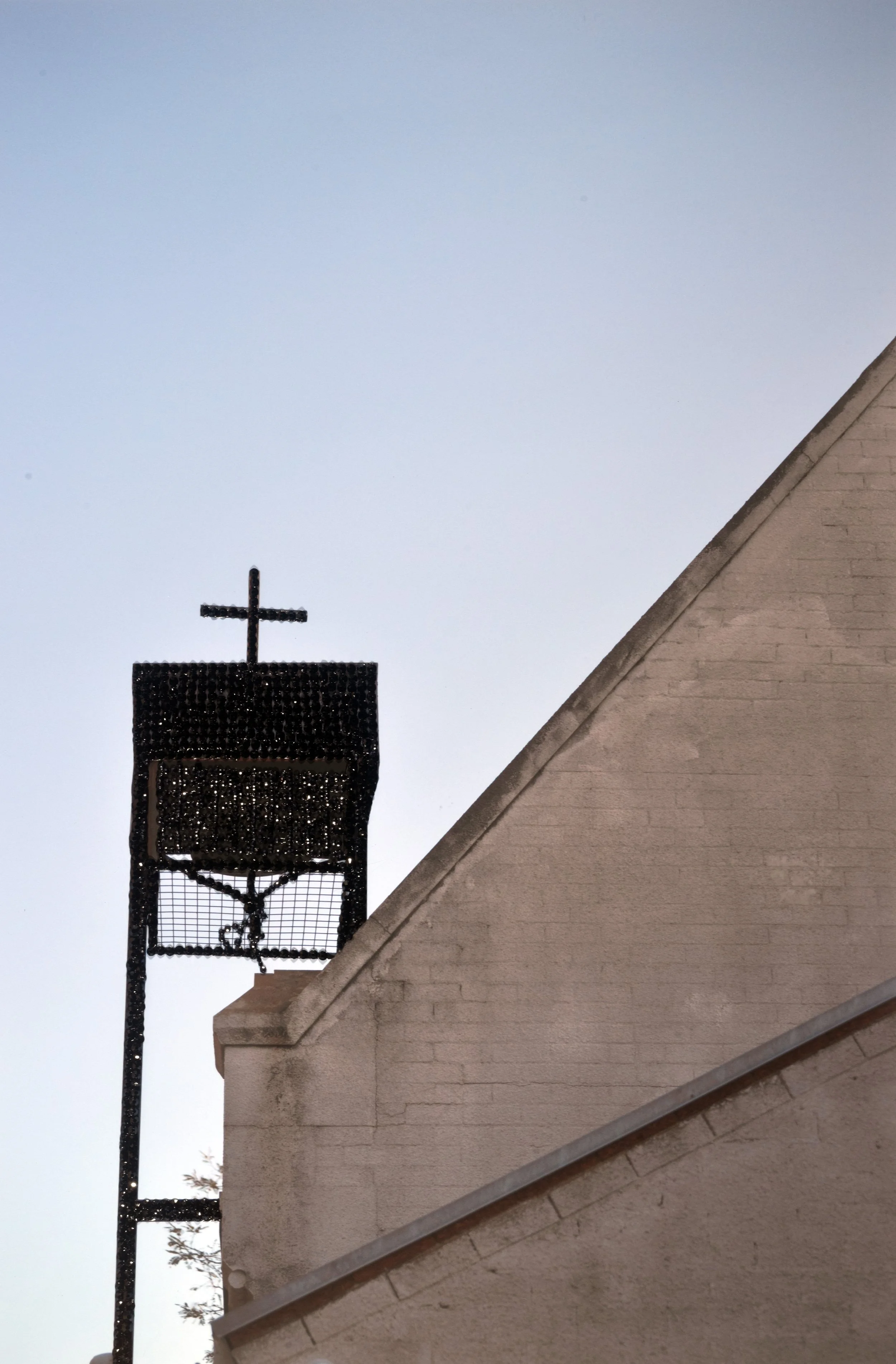 Perth Kintsugi Exhibition- Photograph with beads .A church steeple topped with a cross seen behind a sloped beige brick wall, with a clear blue sky in the background.
