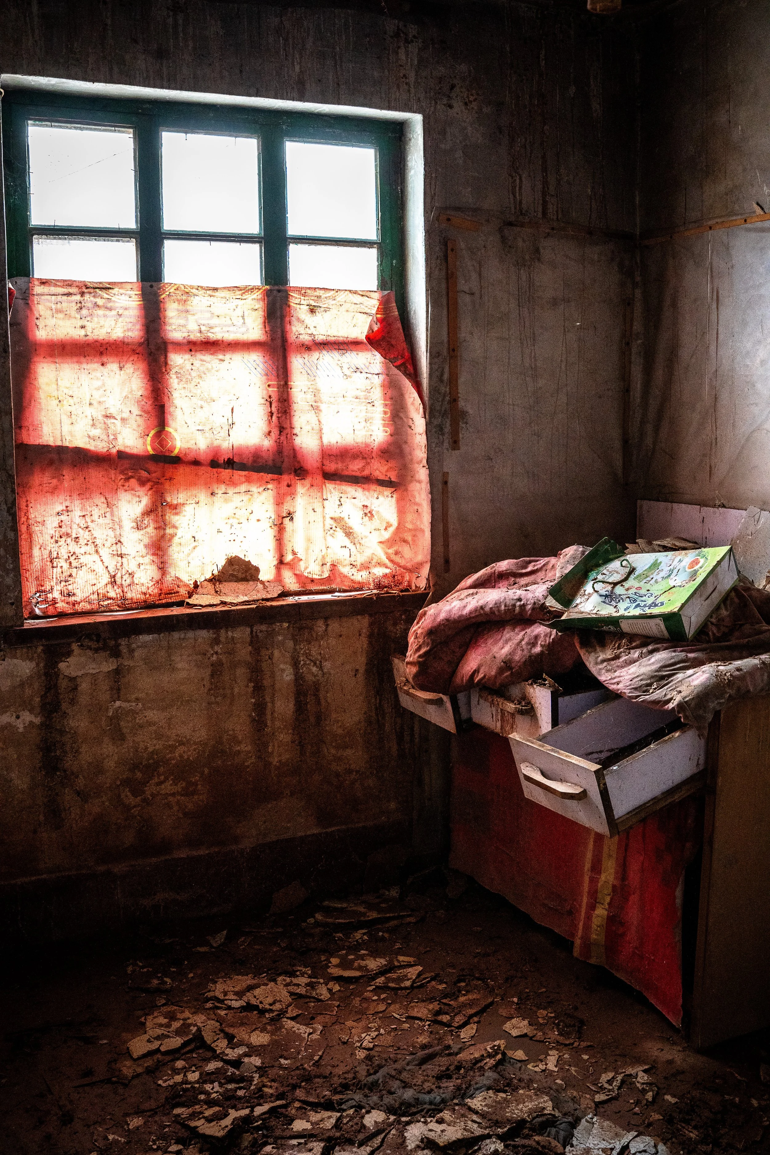 An abandoned room with broken and dusty floor, a window with peeling green paint, and makeshift curtain or cover made of fabric tied in place. There are clutter and what appears to be garbage or debris on a small table or surface on the right side.