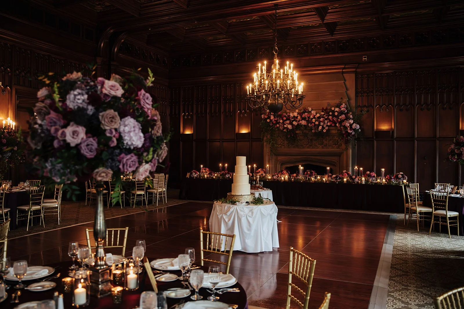 Elegant wedding reception at the University Club of Chicago with dark wood paneling, a large chandelier, floral arrangements, a multi-tiered wedding cake, and decorated tables with candles and gold chairs.