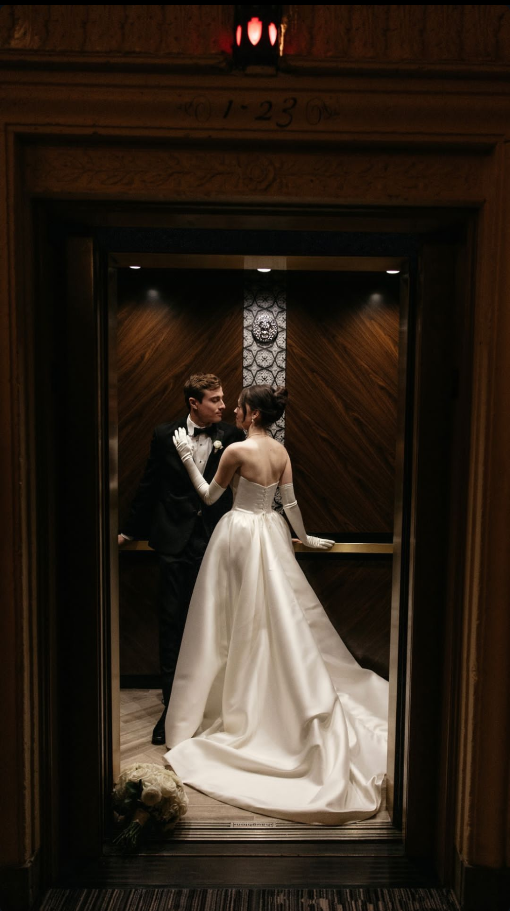 A bride and groom in wedding attire inside an elevator, gazing at each other. The bride wears a strapless white gown with long gloves, and the groom is in a black tuxedo. A bouquet of white flowers is on the floor inside the elevator.