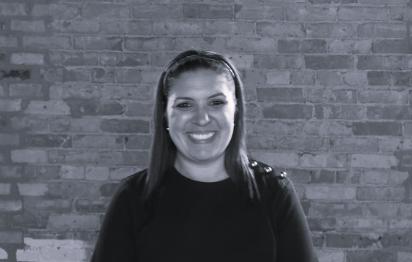 A smiling woman with long hair stands in front of a brick wall, wearing a dark top.