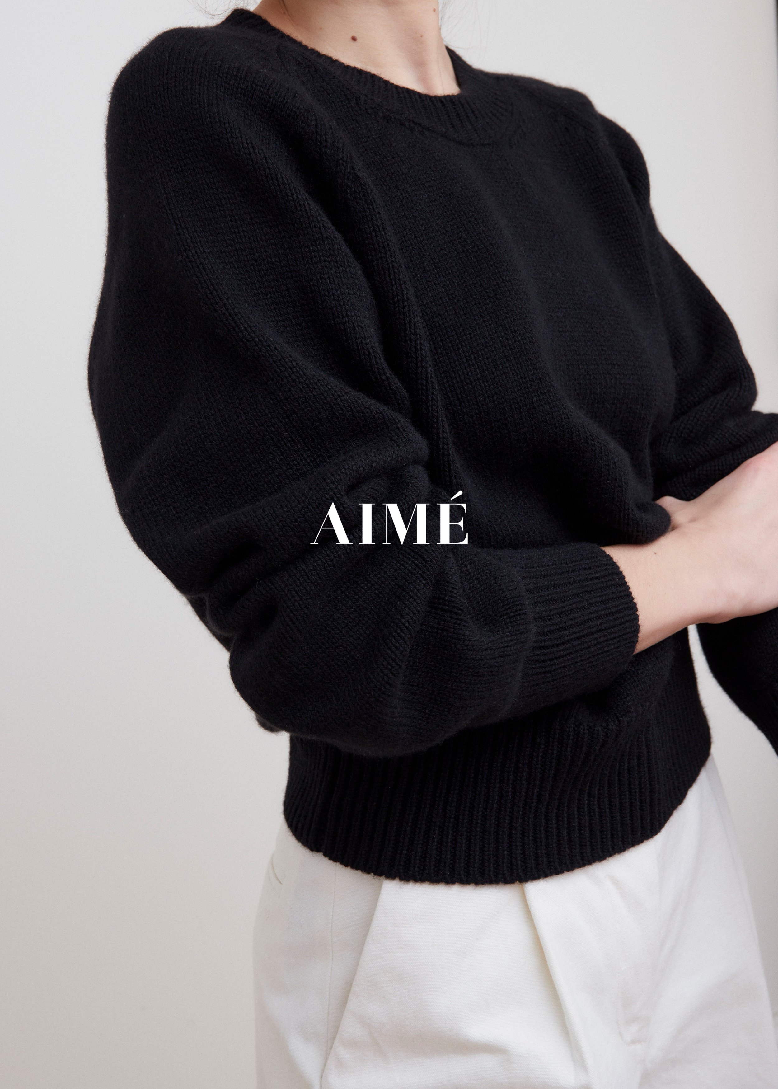 Person wearing a black knit sweater and white pants, with arms crossed, against a plain background.