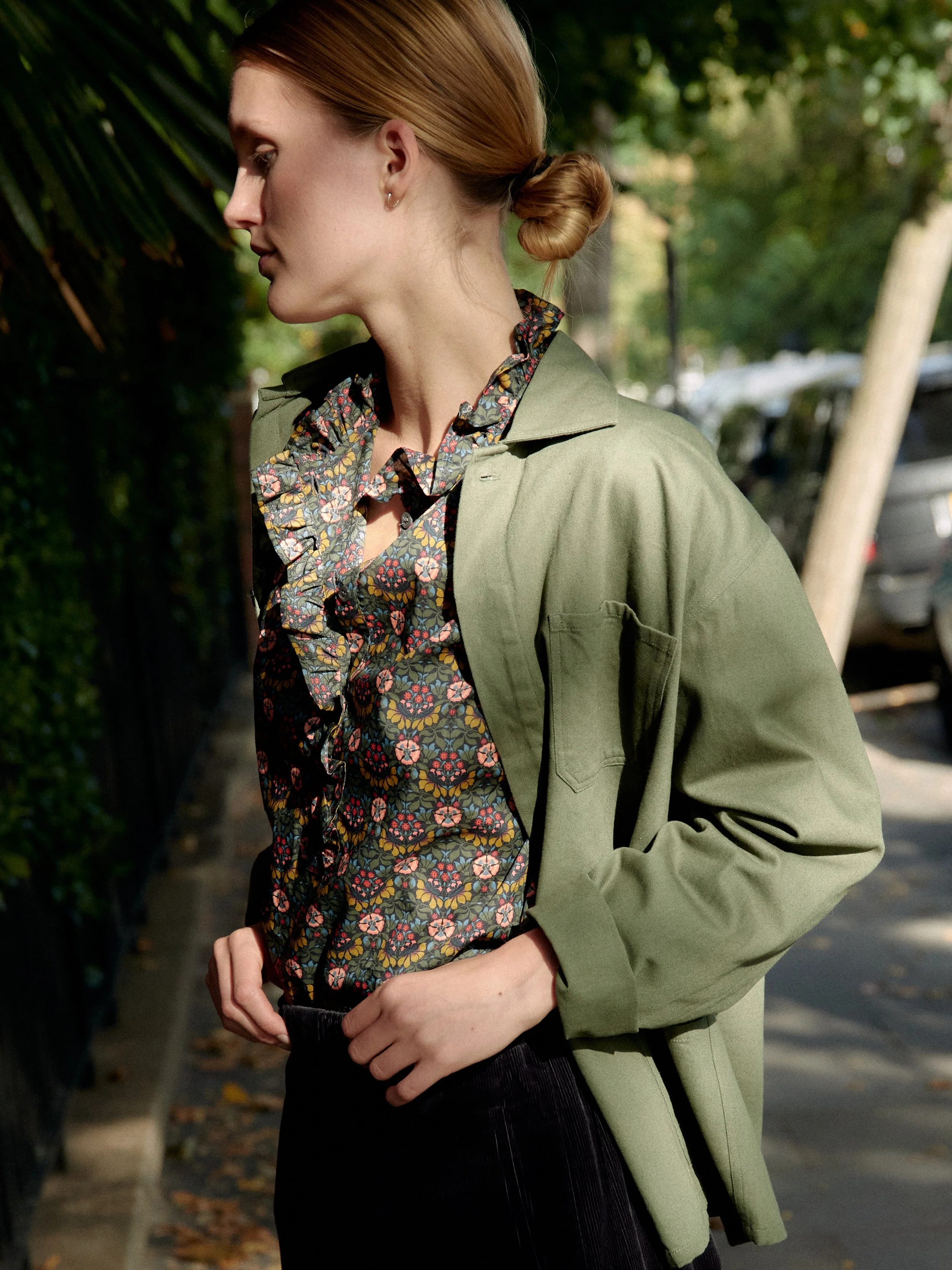 A woman with red hair styled in a bun, wearing a floral-patterned ruffled blouse and a green jacket, standing outdoors on a sidewalk with green trees and parked cars in the background.