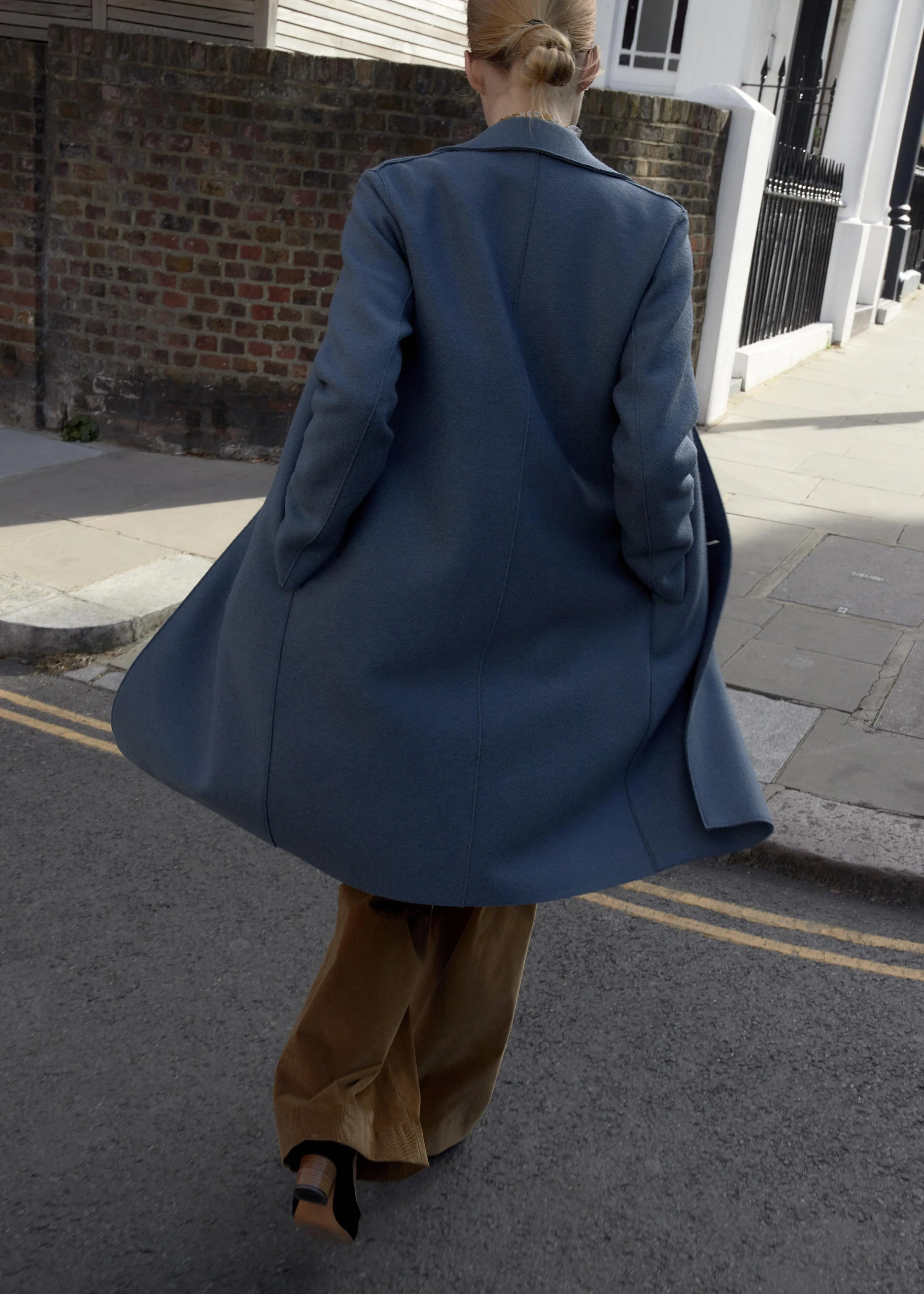 A woman with red hair styled in a bun, wearing a dark blue coat and brown wide-leg pants, walking on a sidewalk with a brick wall and black gate in the background.