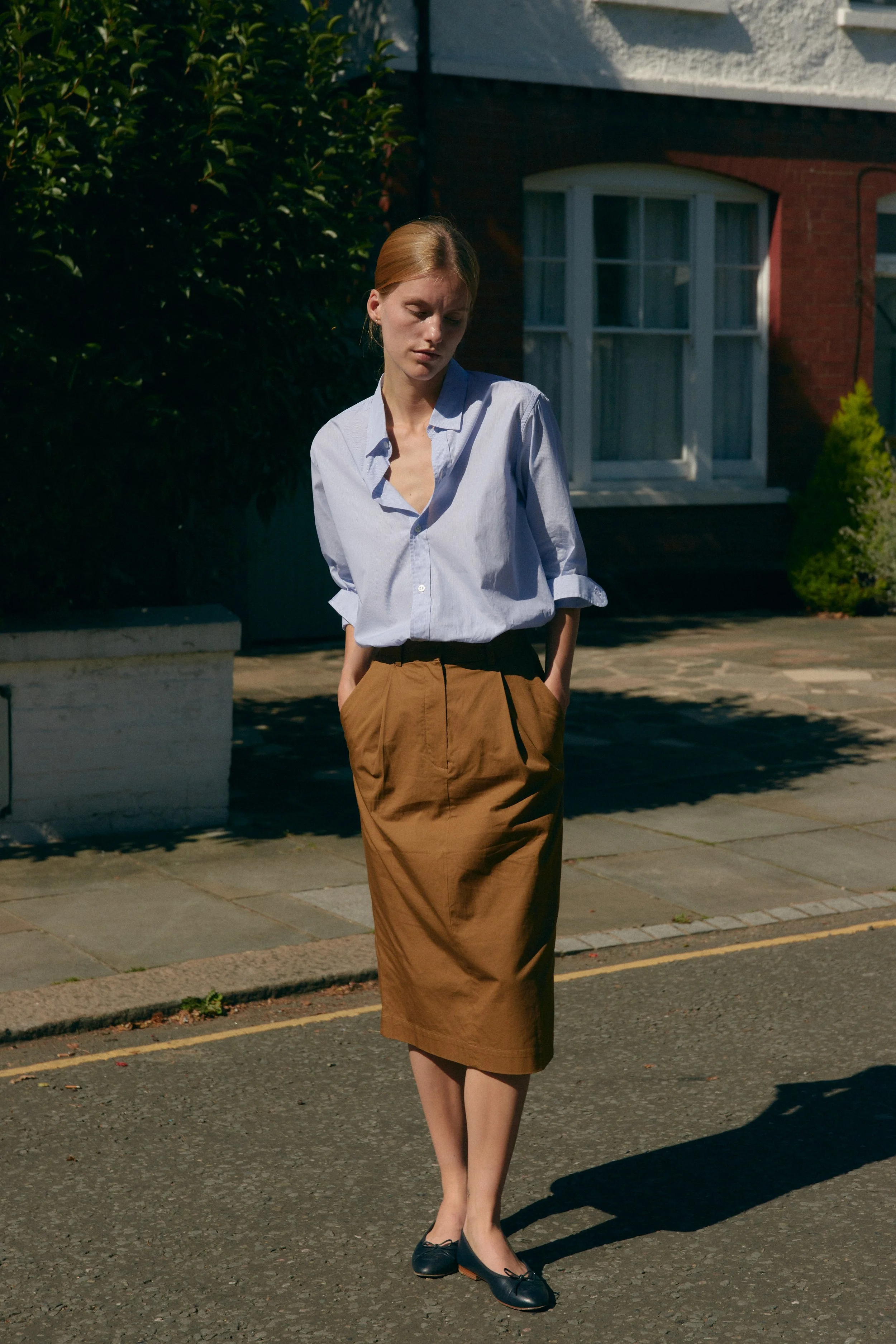 A young woman with reddish hair, dressed in a blue button-up shirt, a brown skirt, and black shoes, walking on a suburban street with her eyes cast downward.